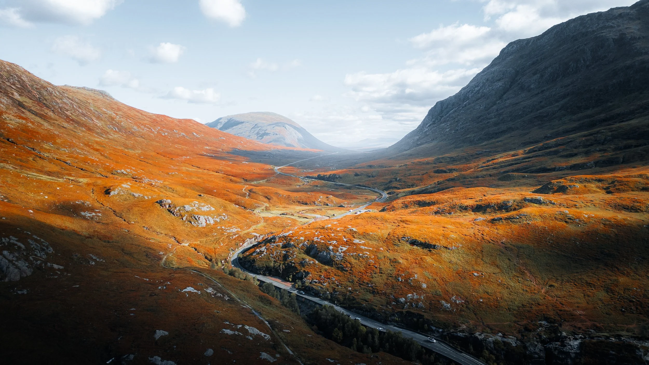 Scenic view of a winding mountain valley with orange and green foliage, steep hills on either side, and a road with cars at the bottom.