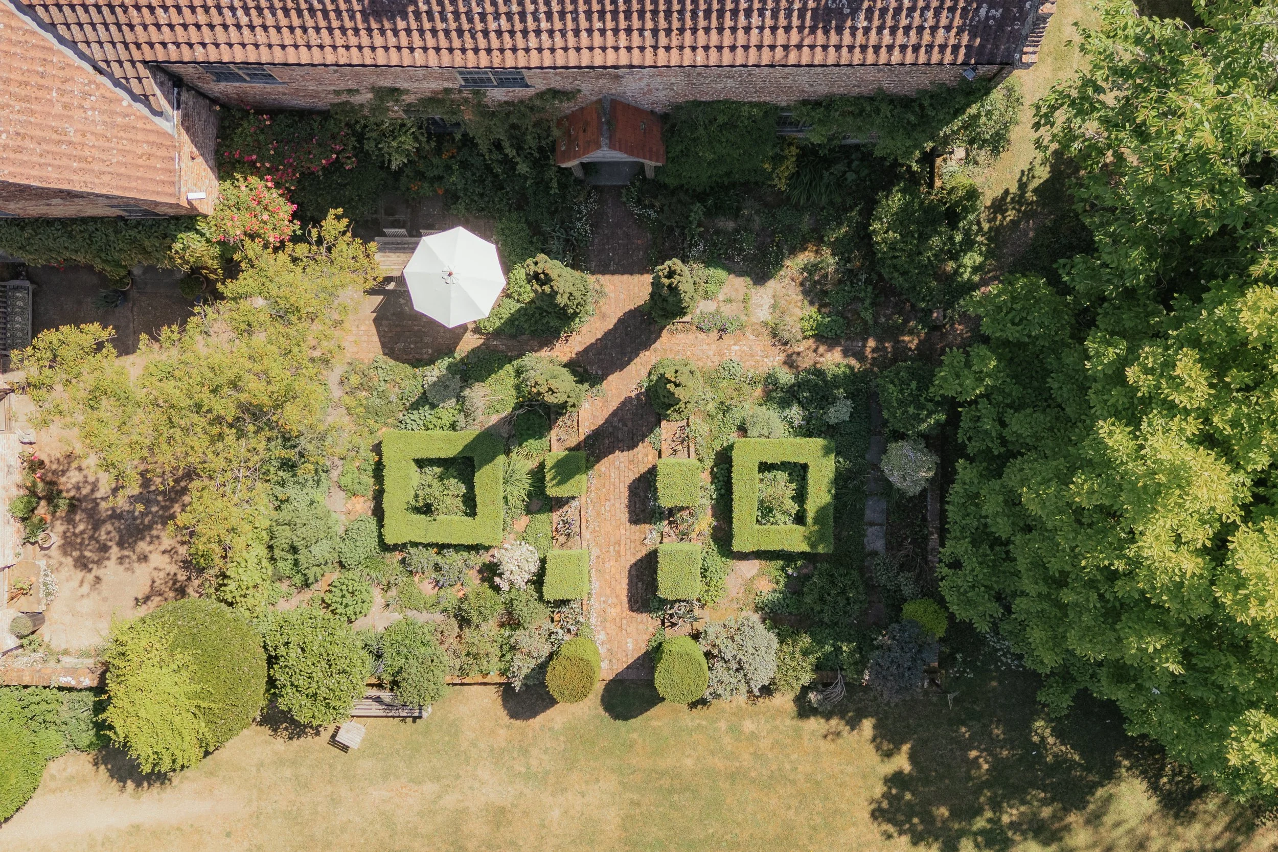 Aerial view of a garden with neatly trimmed bushes, trees, a brick pathway, and a white patio umbrella.