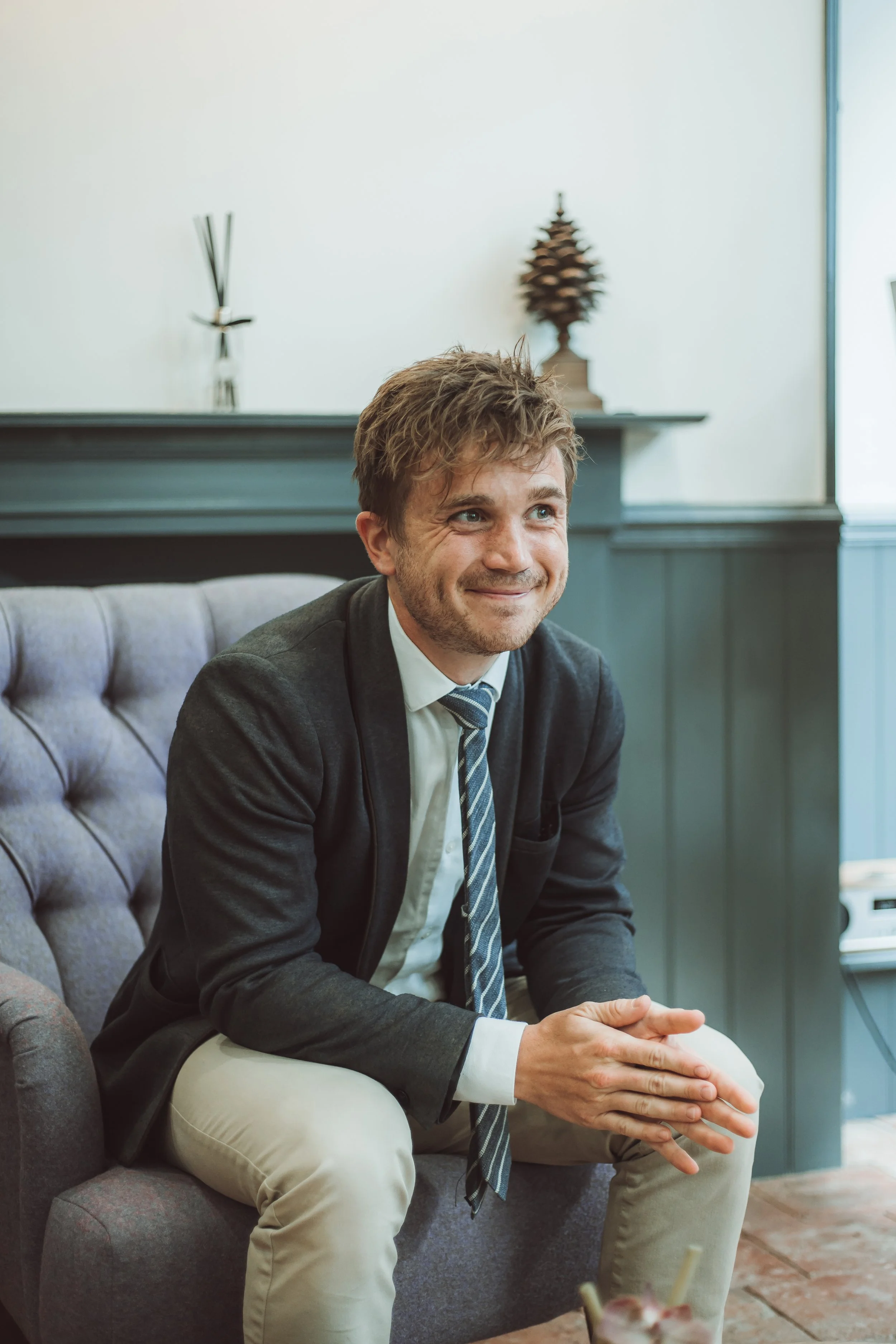 A man in a suit and tie sitting on a sofa, smiling, with a decorative pine cone on the shelf behind him.