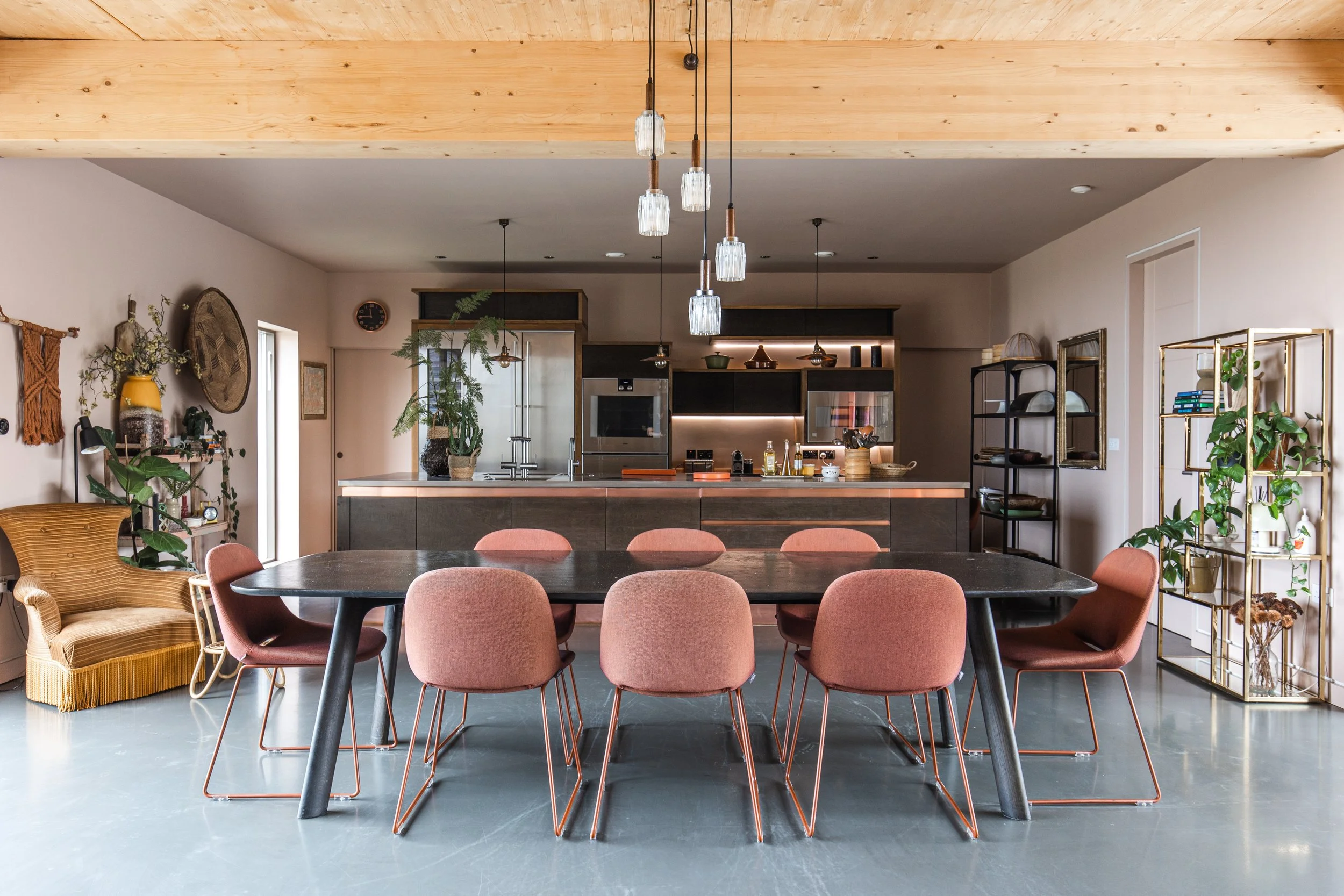 Modern kitchen and dining area with gray flooring, a black table, pink chairs, and a large wooden ceiling beam. Decor includes plants, shelves, and hanging glass pendant lights.