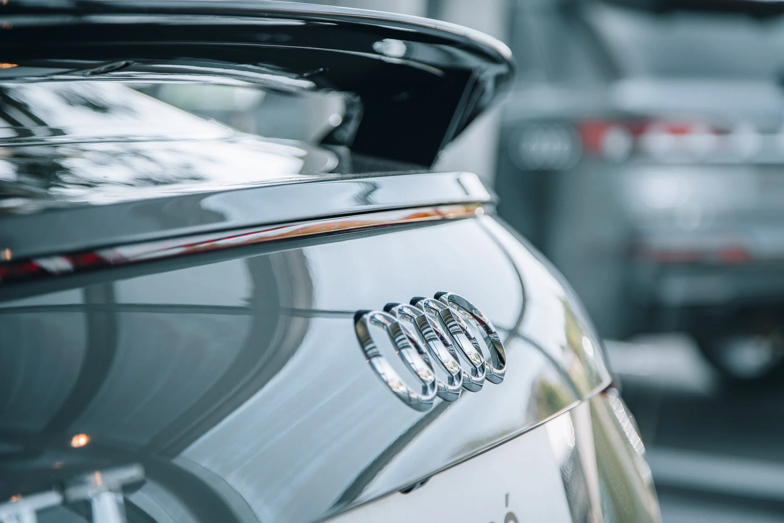 Close-up of the rear of a silver Audi car with the iconic four-ring logo, parked indoors with other cars visible in the background.