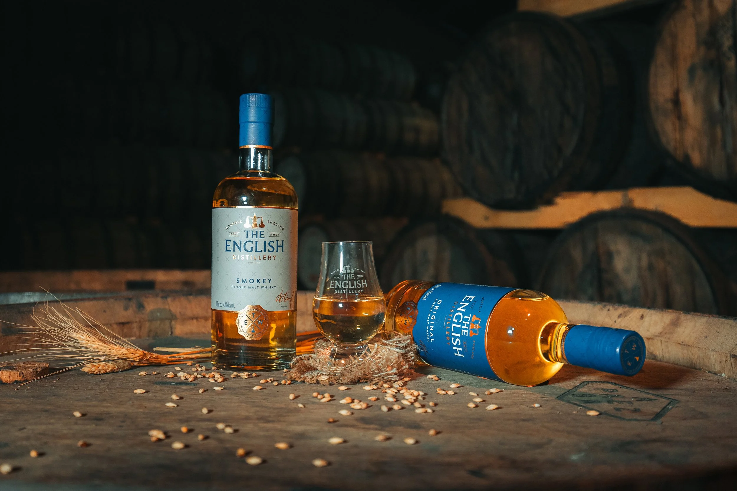 Two bottles of English whisky, one standing upright and one lying down, a glass of whisky, some wheat, and scattered grains on a wooden surface in a barrel storage setting.
