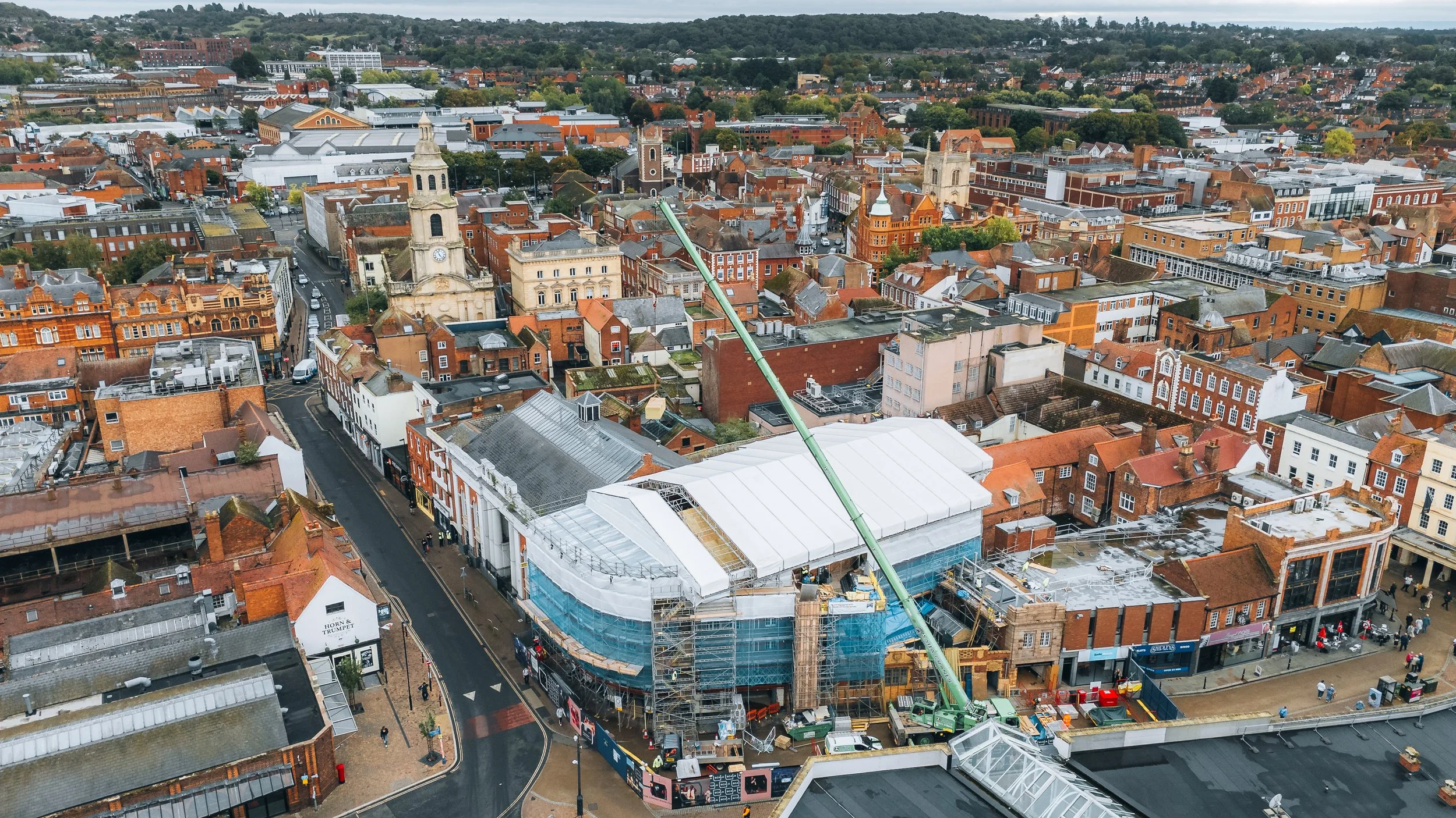 Aerial view of a city with a mix of historic and modern buildings, with a green crane and construction scaffolding on a large building in the foreground.