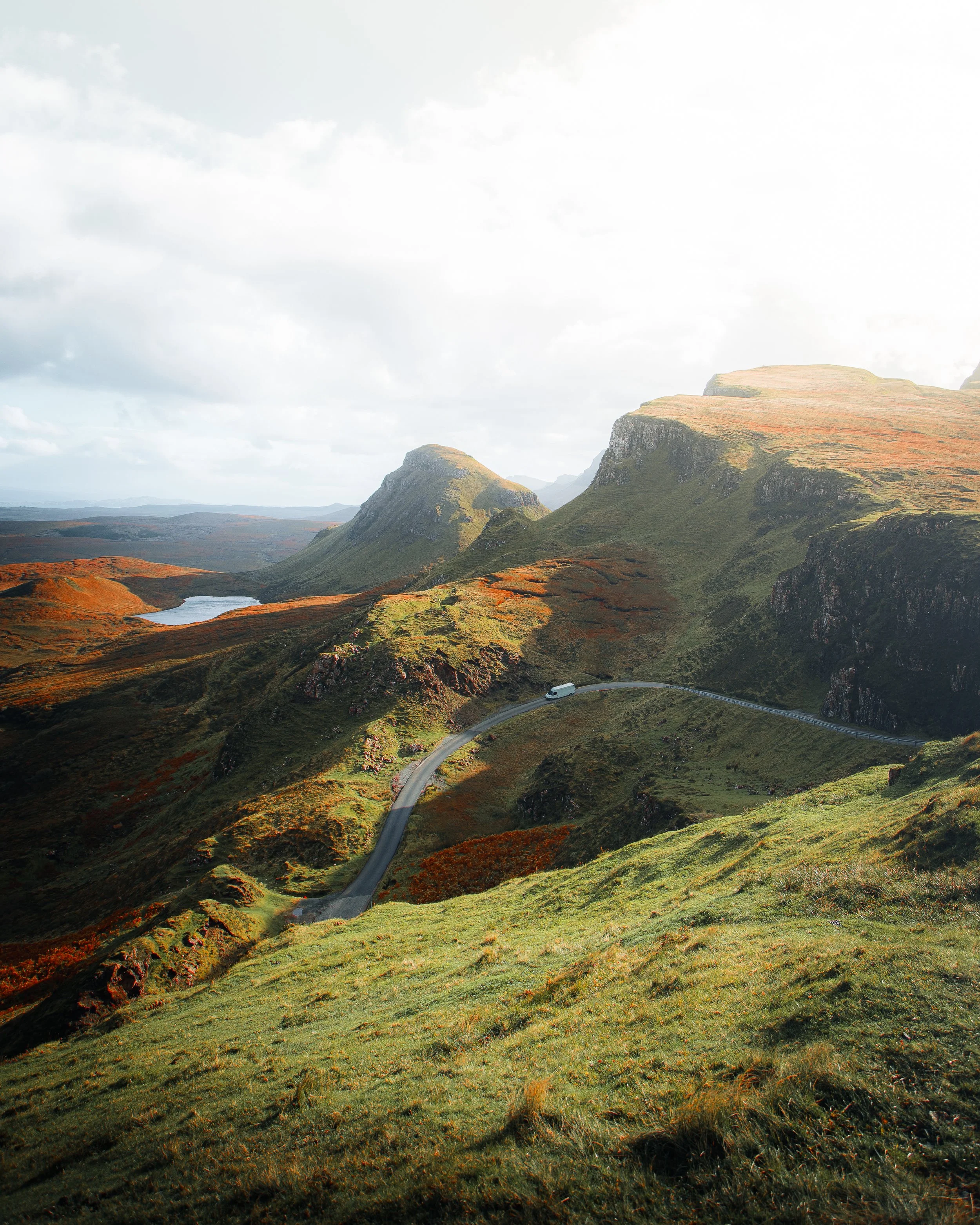 Scenic view of green, orange, and brown mountains with a winding road, a white van, and a small lake, under a partly cloudy sky.