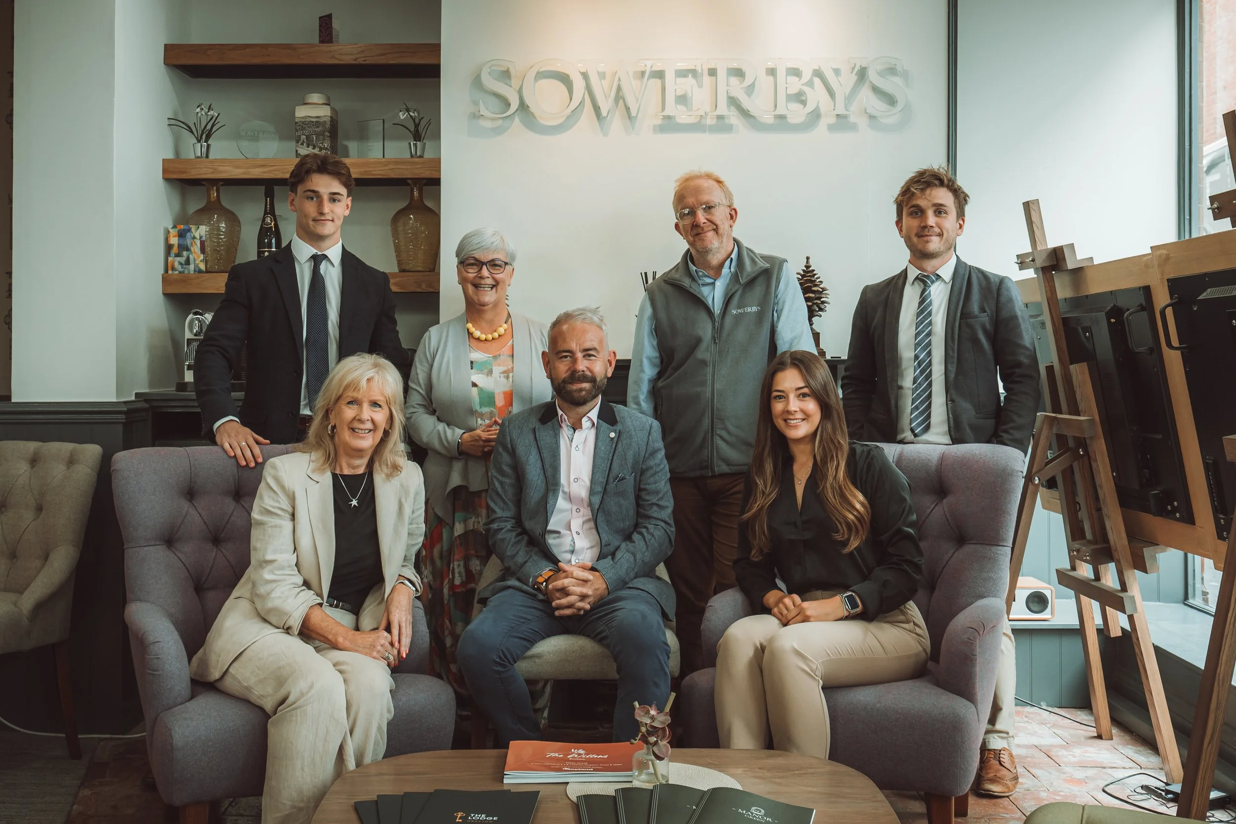 Group of eight people, including both men and women of various ages, pose for a photo in a well-lit, modern office or lounge area with a sign that reads 'SOWERBYS' on the wall behind them. They are dressed in business casual or professional attire, w