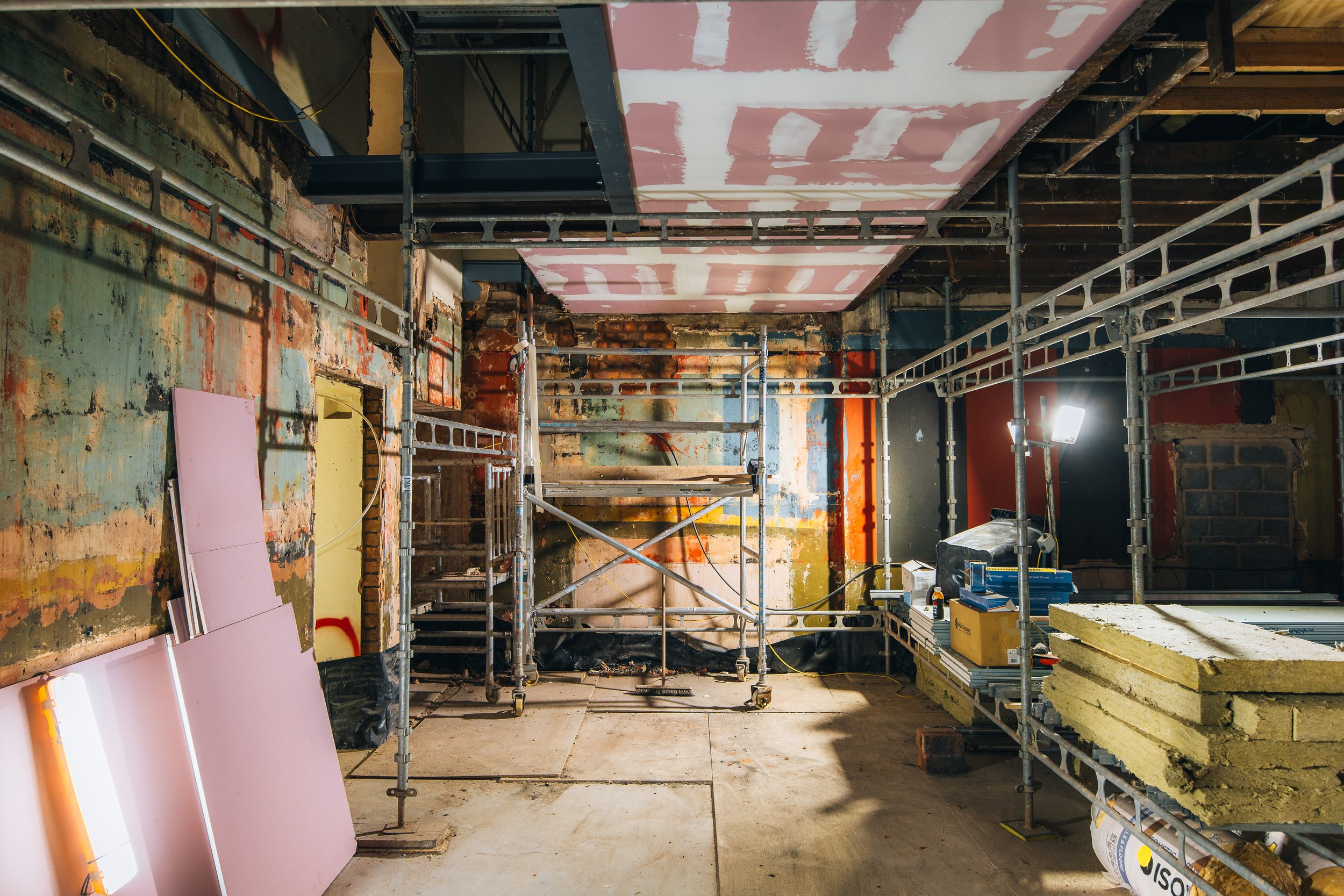 Interior of a building under renovation with scaffolding, exposed brick and drywall, construction materials, and tools.