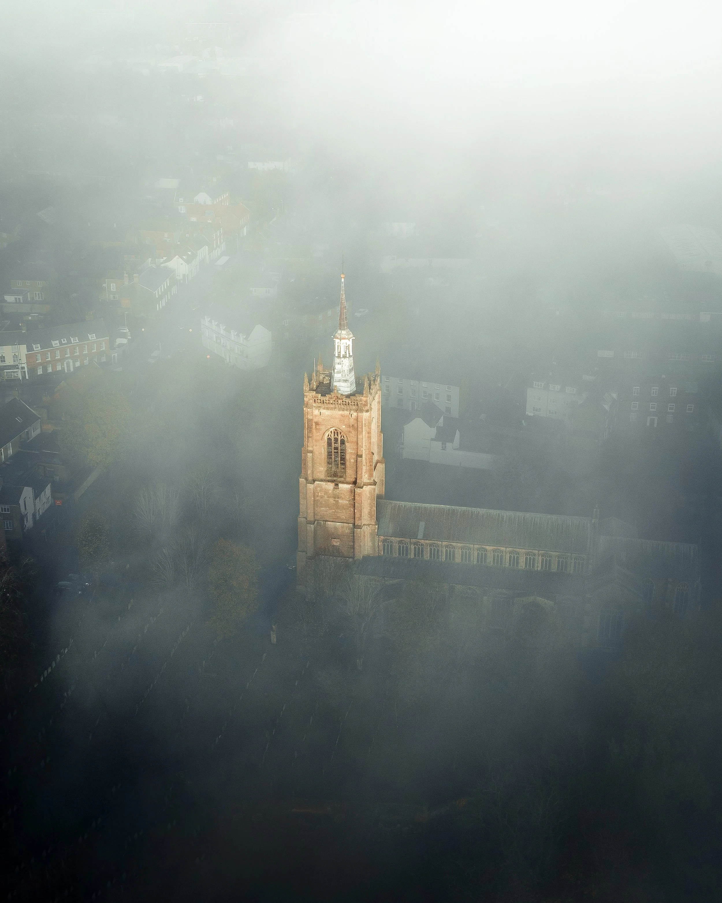 Aerial view of a church surrounded by fog, with a tall spire and a graveyard in the foreground.