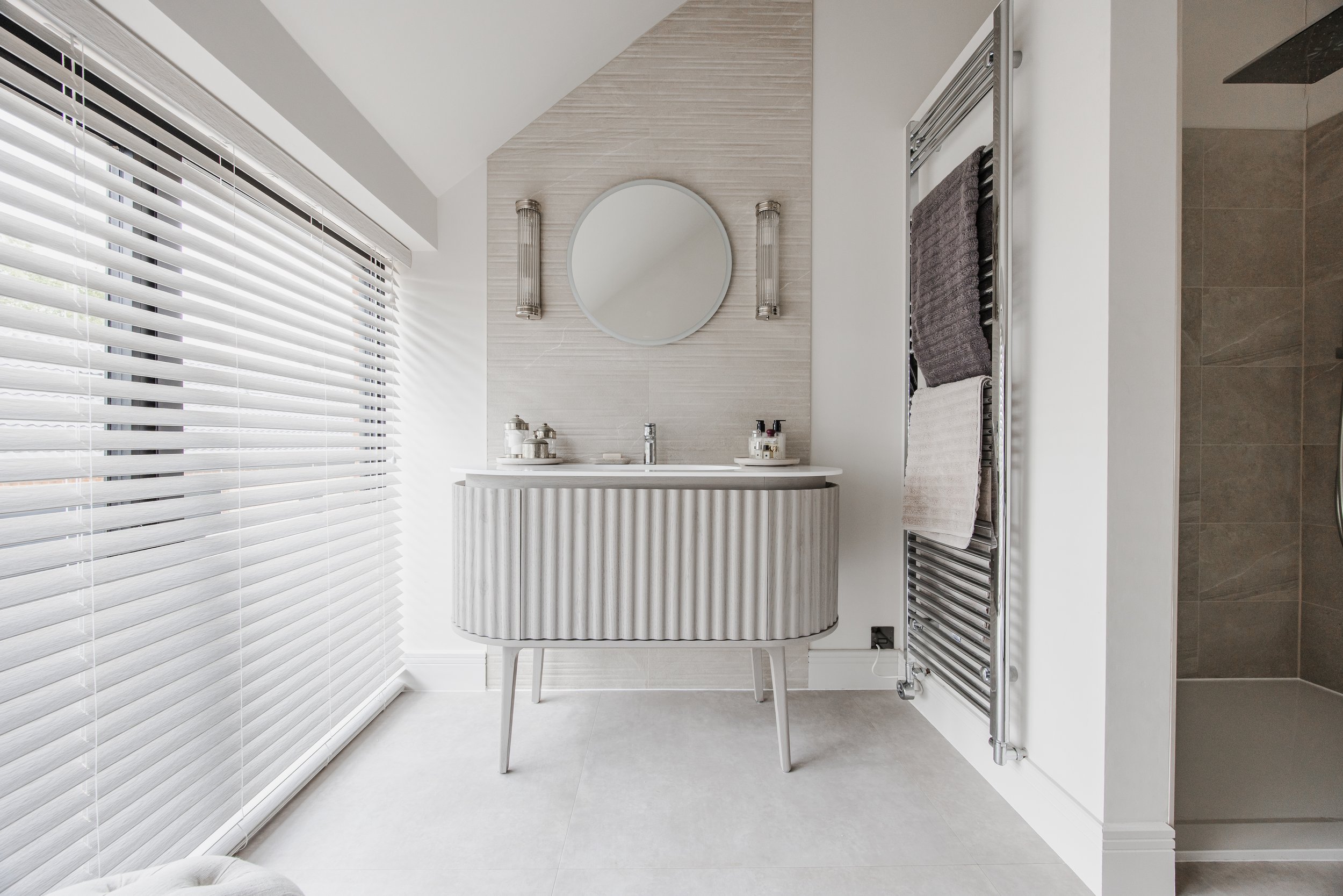 Modern bathroom with a white sink and mirror, gray towels on a heated towel rail, and a walk-in shower with brown tiles.