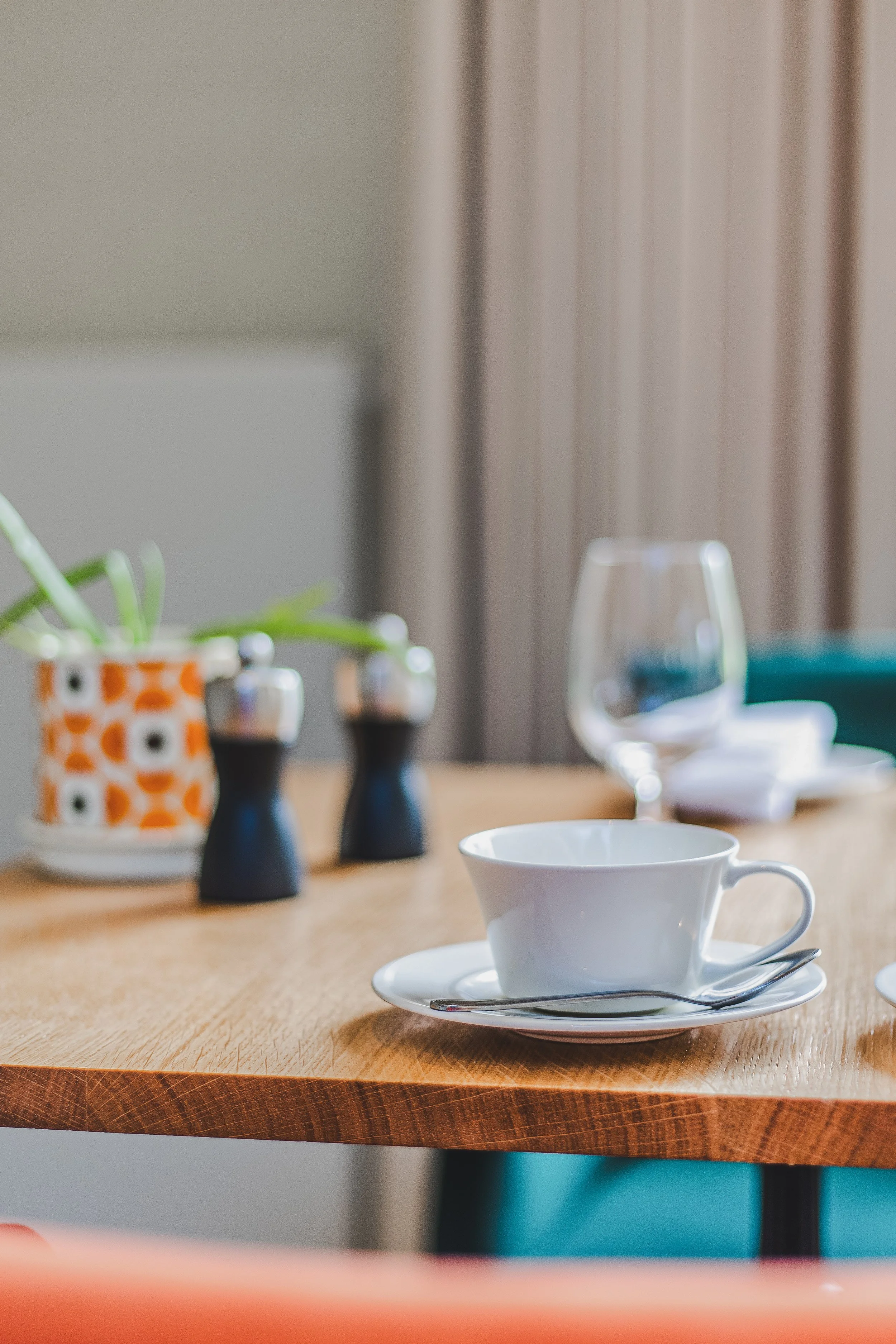 A close-up of a white teacup on a saucer with a small spoon, placed on a wooden table, with a blurred background showing a wine glass, salt and pepper shakers, a small potted plant, and a teal chair.
