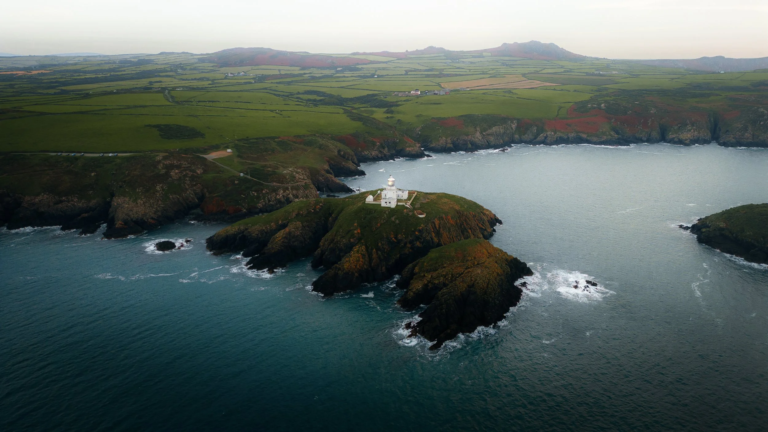 An aerial view of a lighthouse on a rocky island surrounded by the ocean with green rolling hills and farmland in the background.