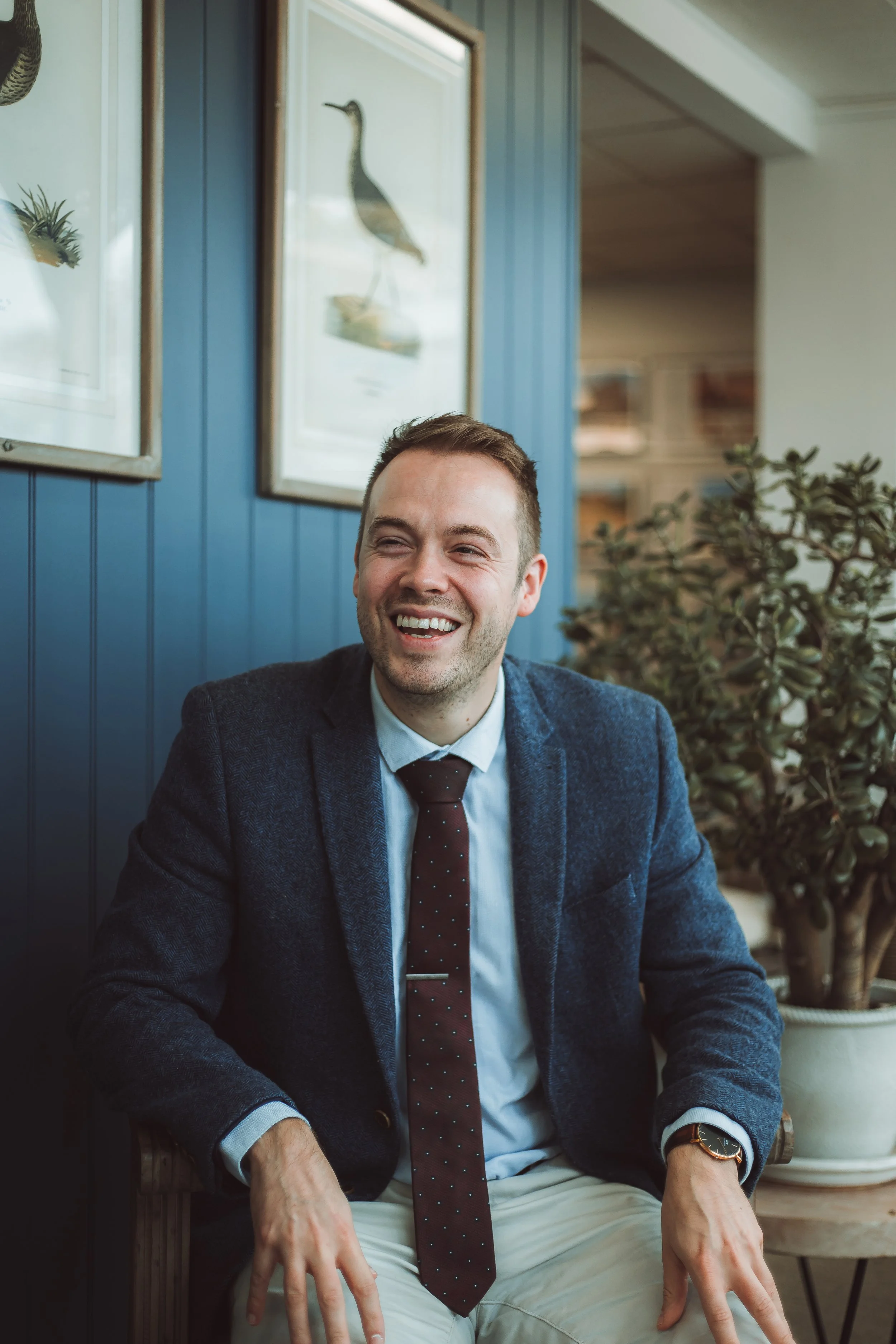 A man sitting indoors, smiling and wearing a dark blue blazer, light blue shirt, dark tie, beige pants, and a watch, with framed bird illustrations on the wall behind him.
