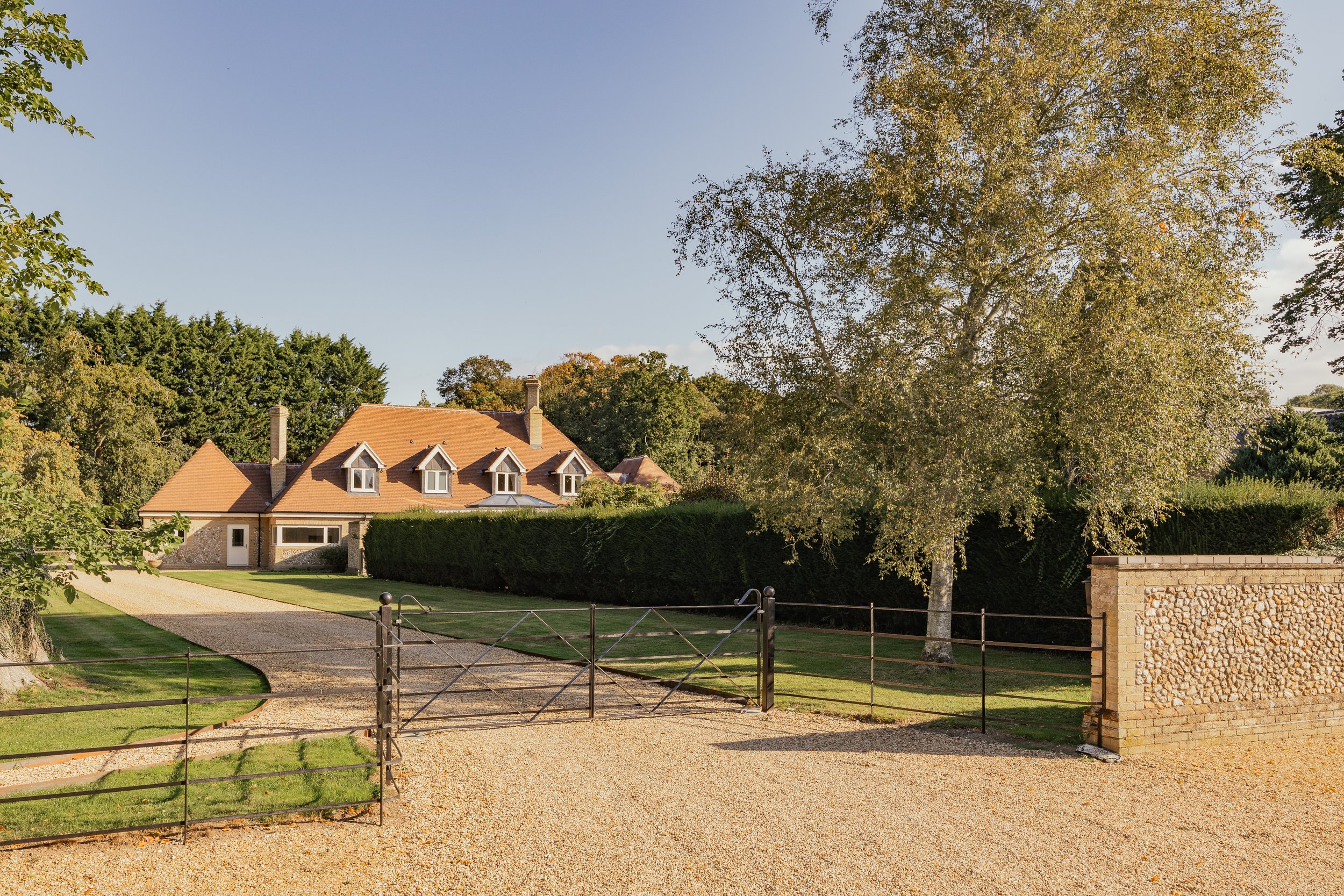 A large house with a red roof, surrounded by green trees and a manicured lawn, enclosed by a black metal gate and gravel driveway.