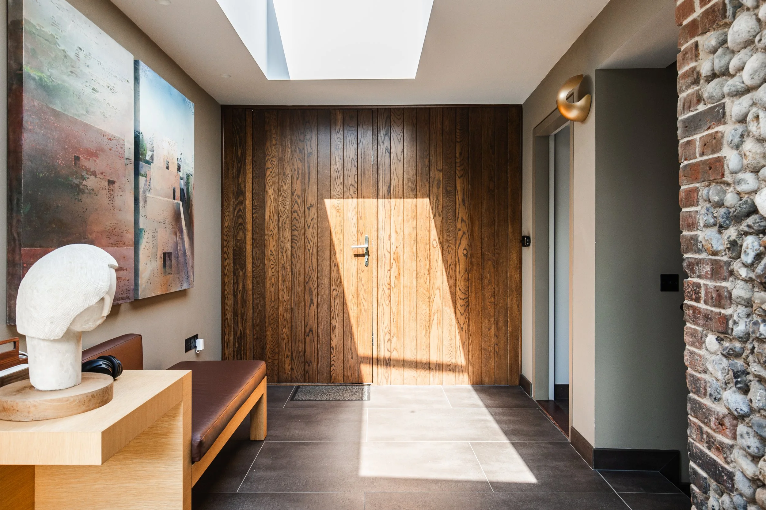 Modern indoor entryway with a wooden door, abstract paintings on the wall, a bench, a sculpture of a head, and a skylight letting in sunlight.