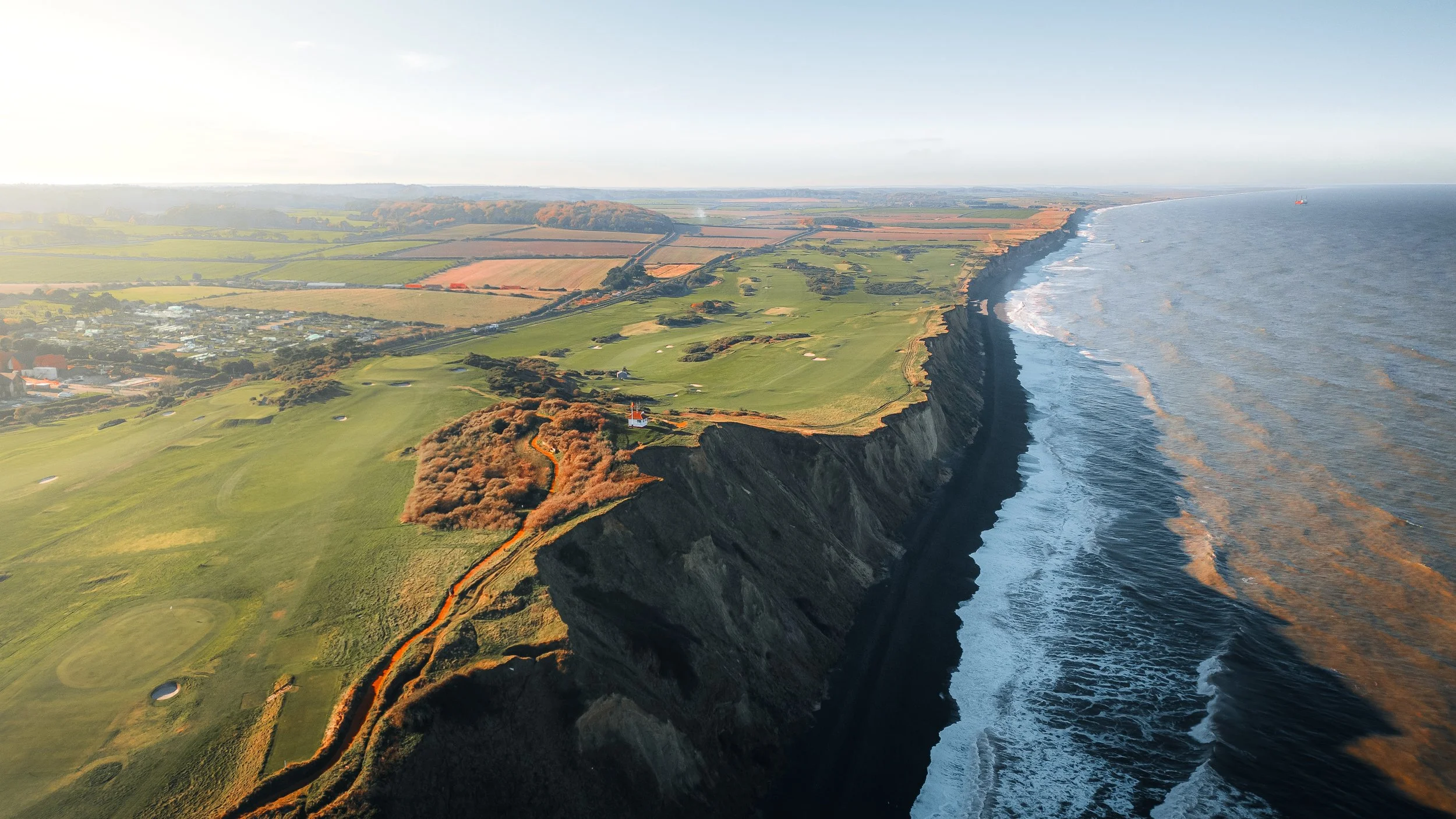 Aerial view of rugged coastal cliffs with green fields and a golf course, overlooking the ocean with waves crashing at the base.