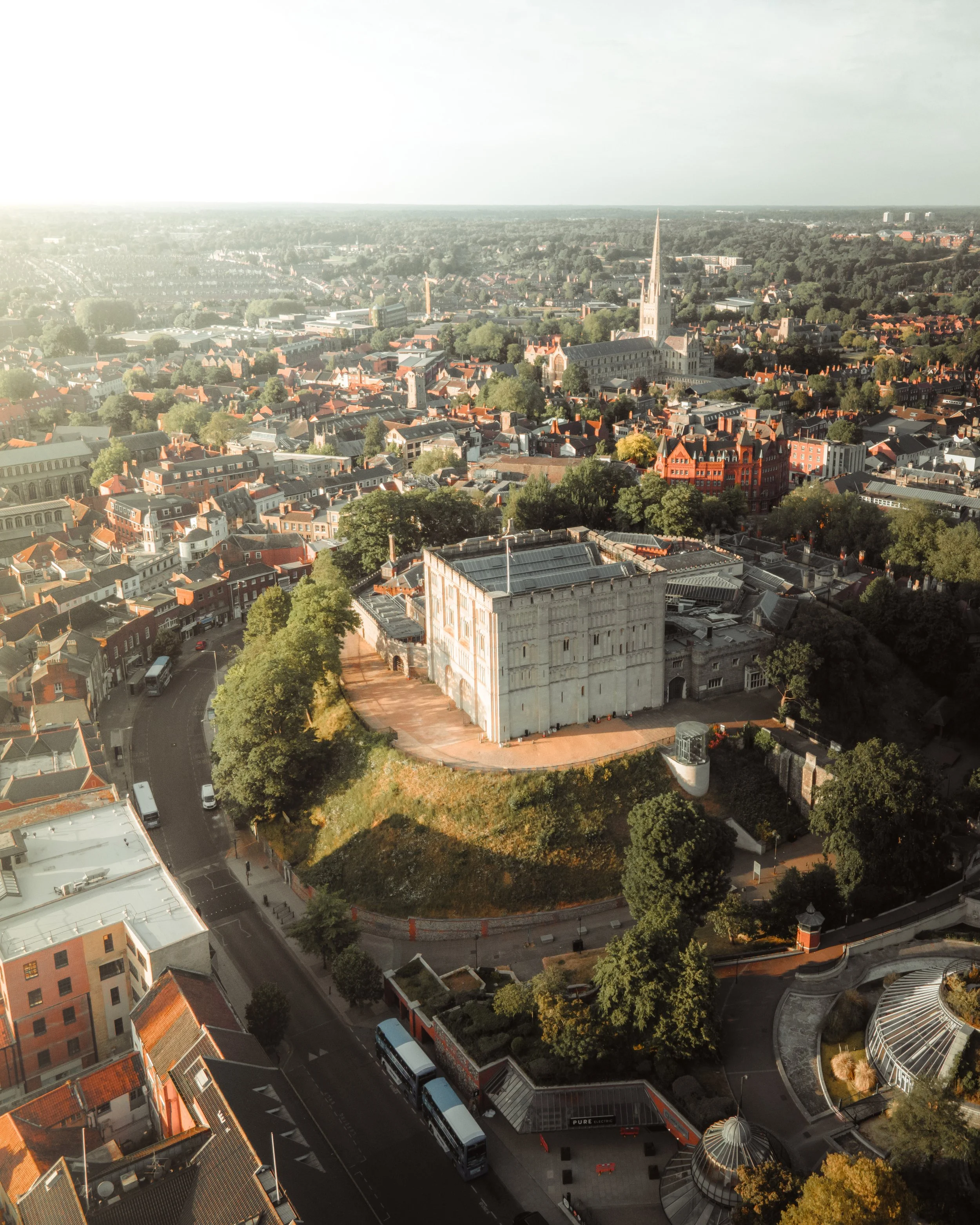 Aerial view of a historic city with a prominent castle on a hill, surrounded by a mix of modern and traditional buildings, streets, trees, and a church with a tall steeple.