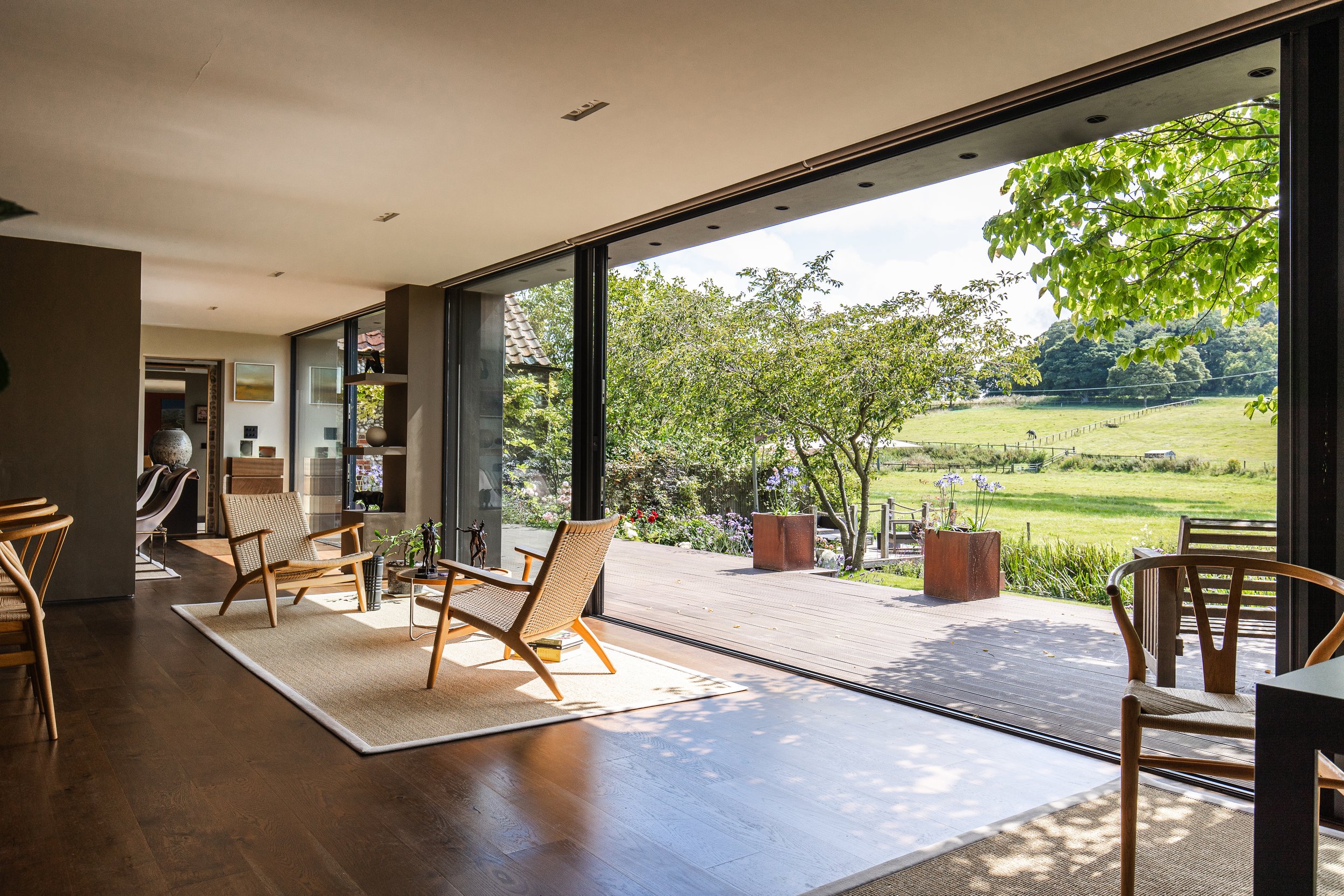 Living room with large glass sliding doors opening to a garden and pasture, with wooden chairs, a white rug, and decorative objects.