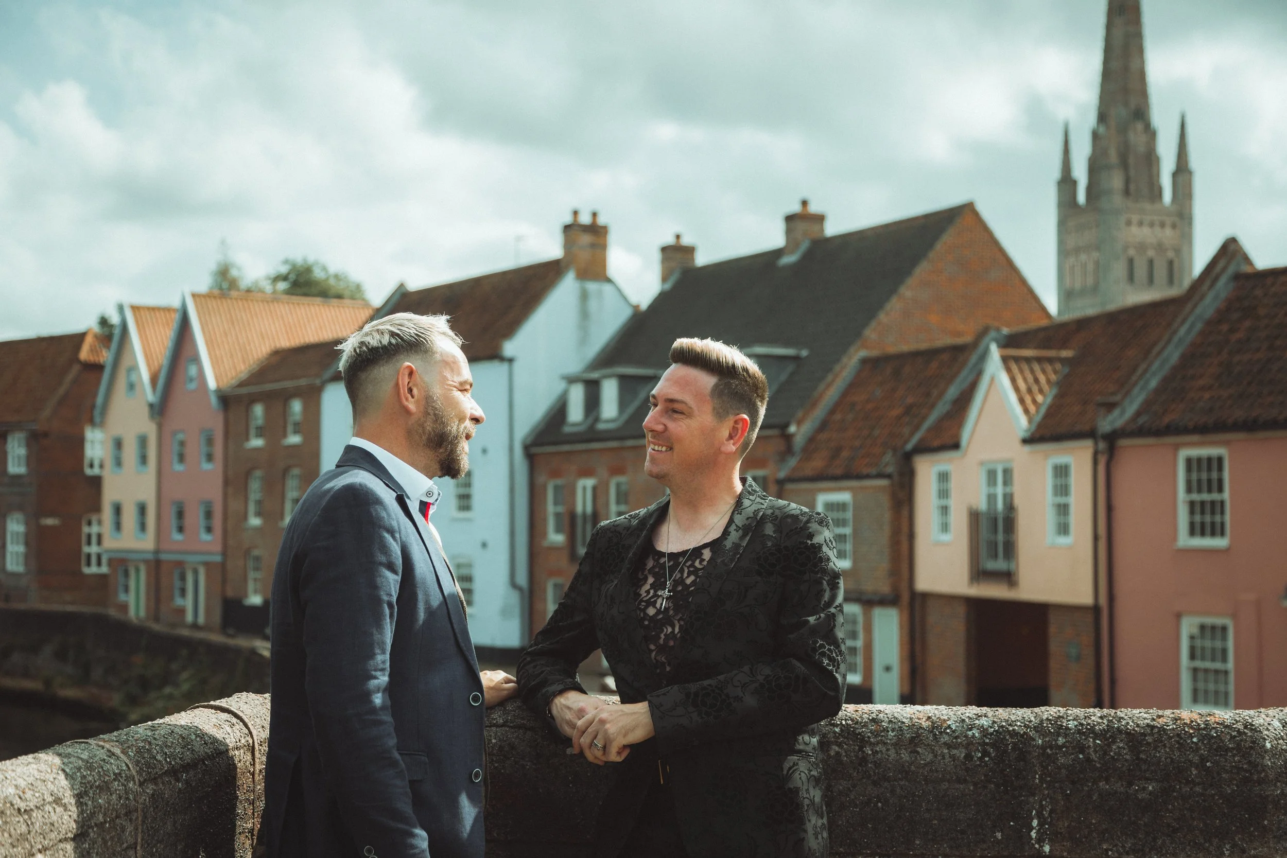 Two men smiling and talking on a bridge in a charming town with colorful houses and a church steeple in the background.