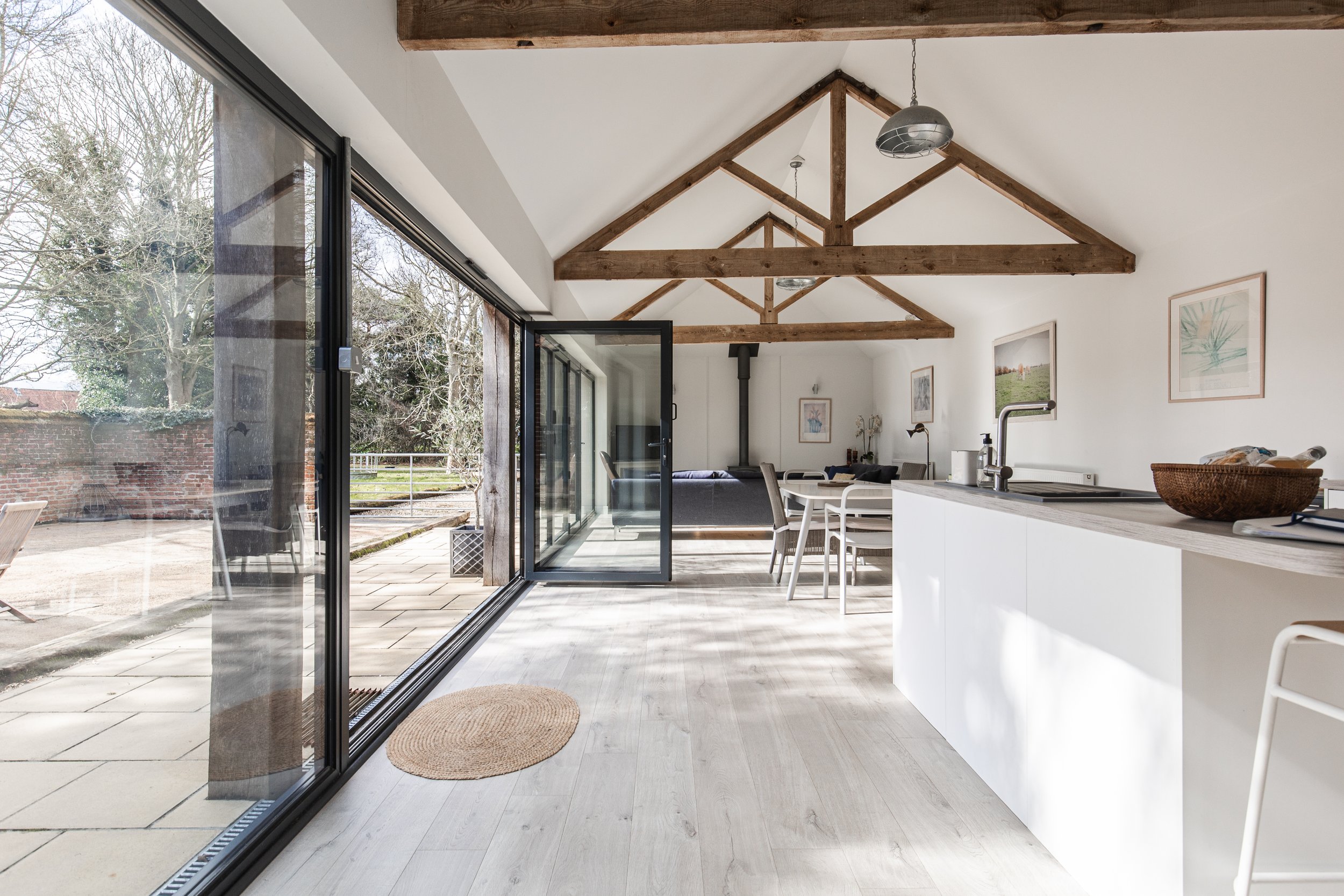 Open-concept living and dining area with a farmhouse kitchen, white walls, exposed wooden beams, large glass sliding doors leading to a patio, and minimal decor.