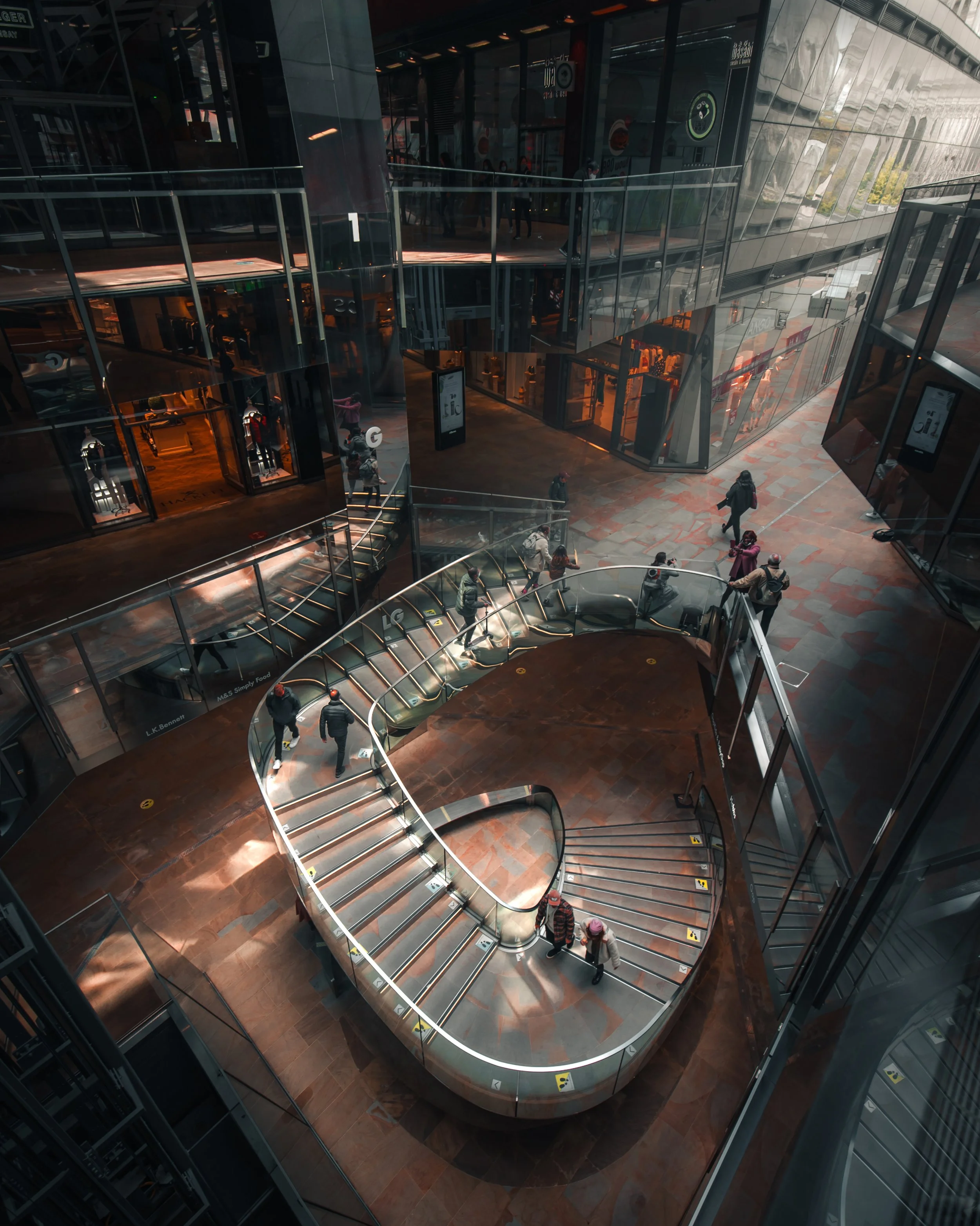 Interior view of a modern, multi-level shopping mall with glass walls, escalators, and shoppers walking on the floors.