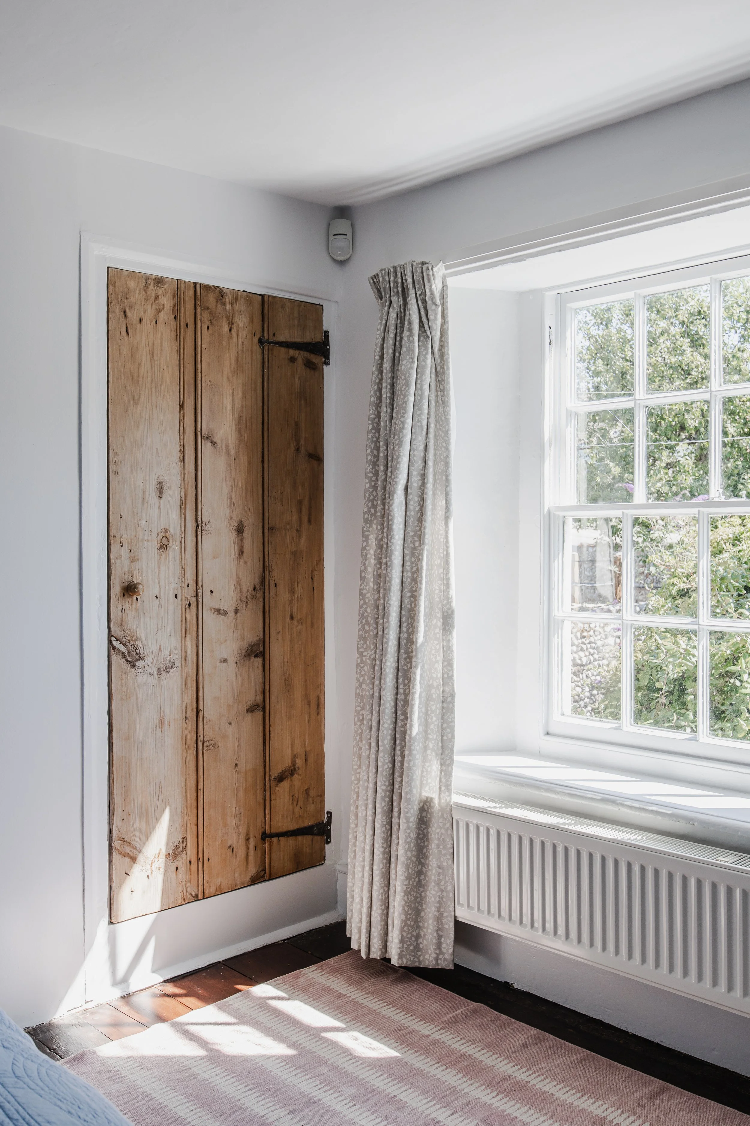 A corner of a room showing a wooden door, a window with curtains, sunlight streaming in, a painted radiator, and a striped rug.