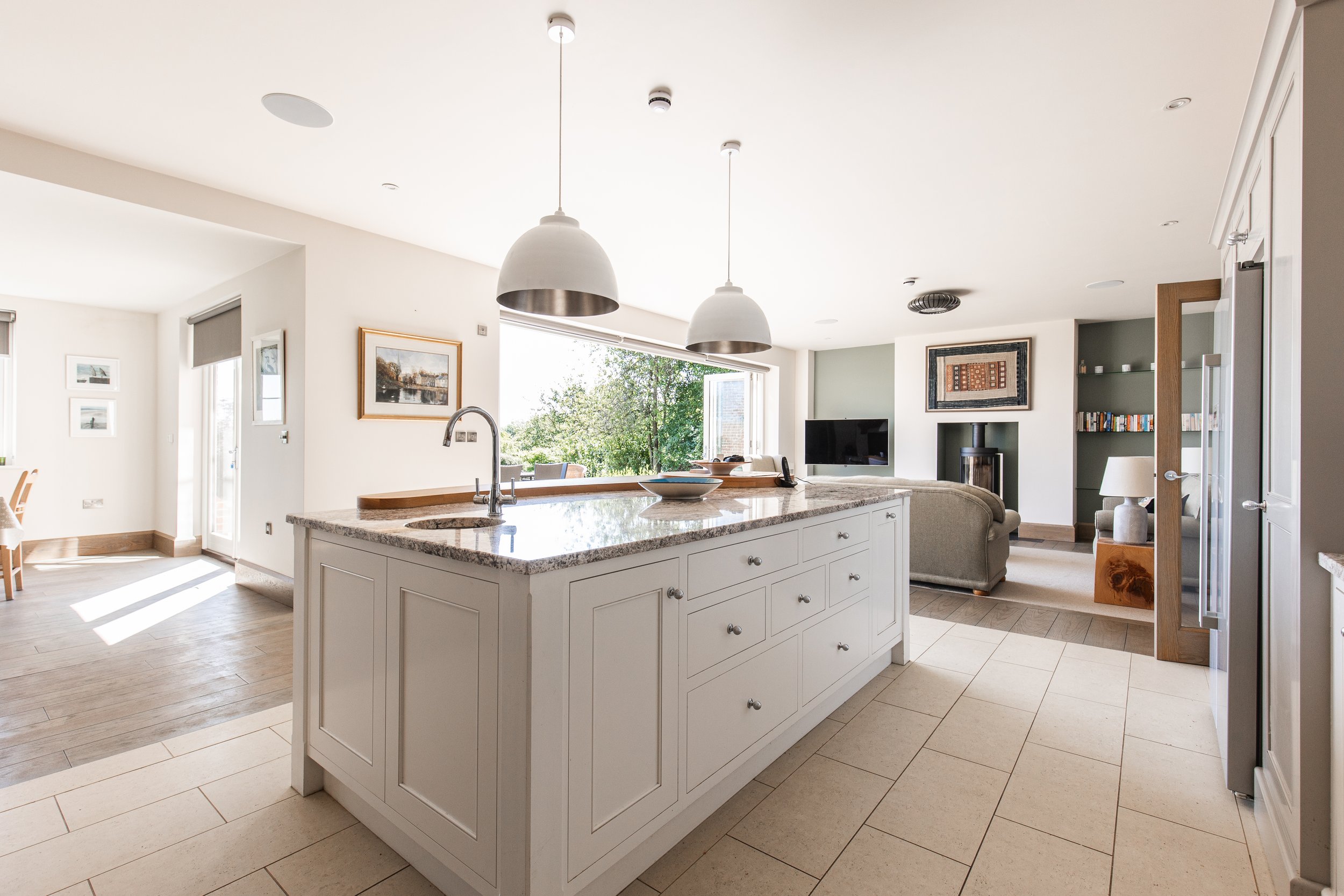 Bright open-concept kitchen and living room with white walls, granite island, pendant lights, and large window showing greenery outside.