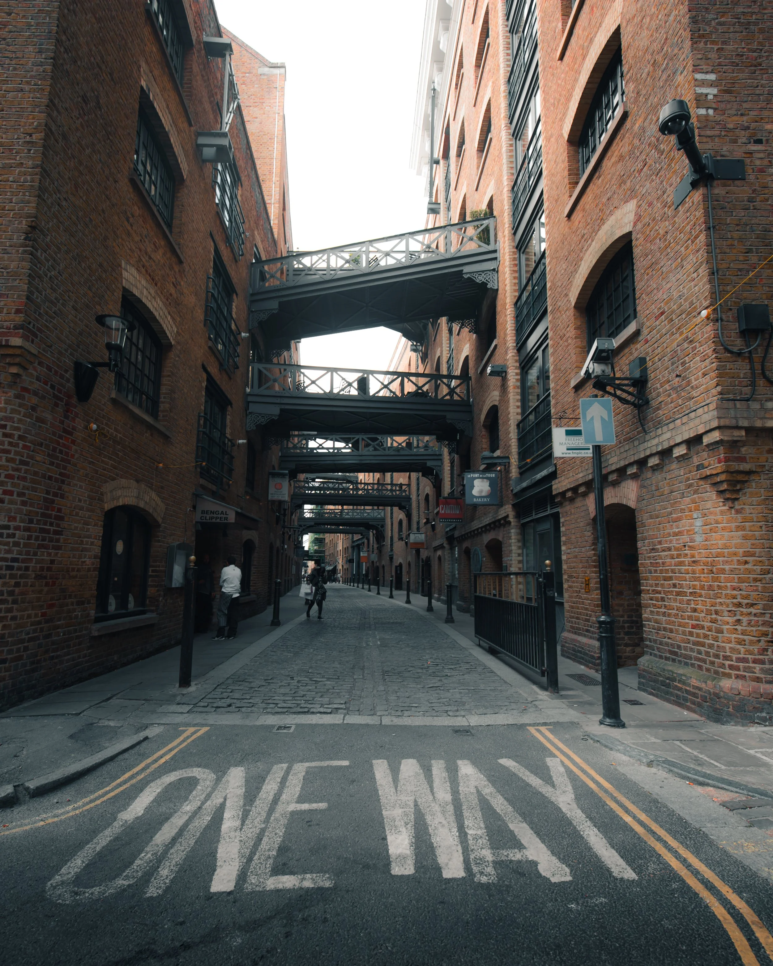 View of a narrow, brick alleyway with cobblestone street, connected by several black metal bridges between buildings. One-way street markings are visible on the road, with pedestrians walking along the sidewalk.