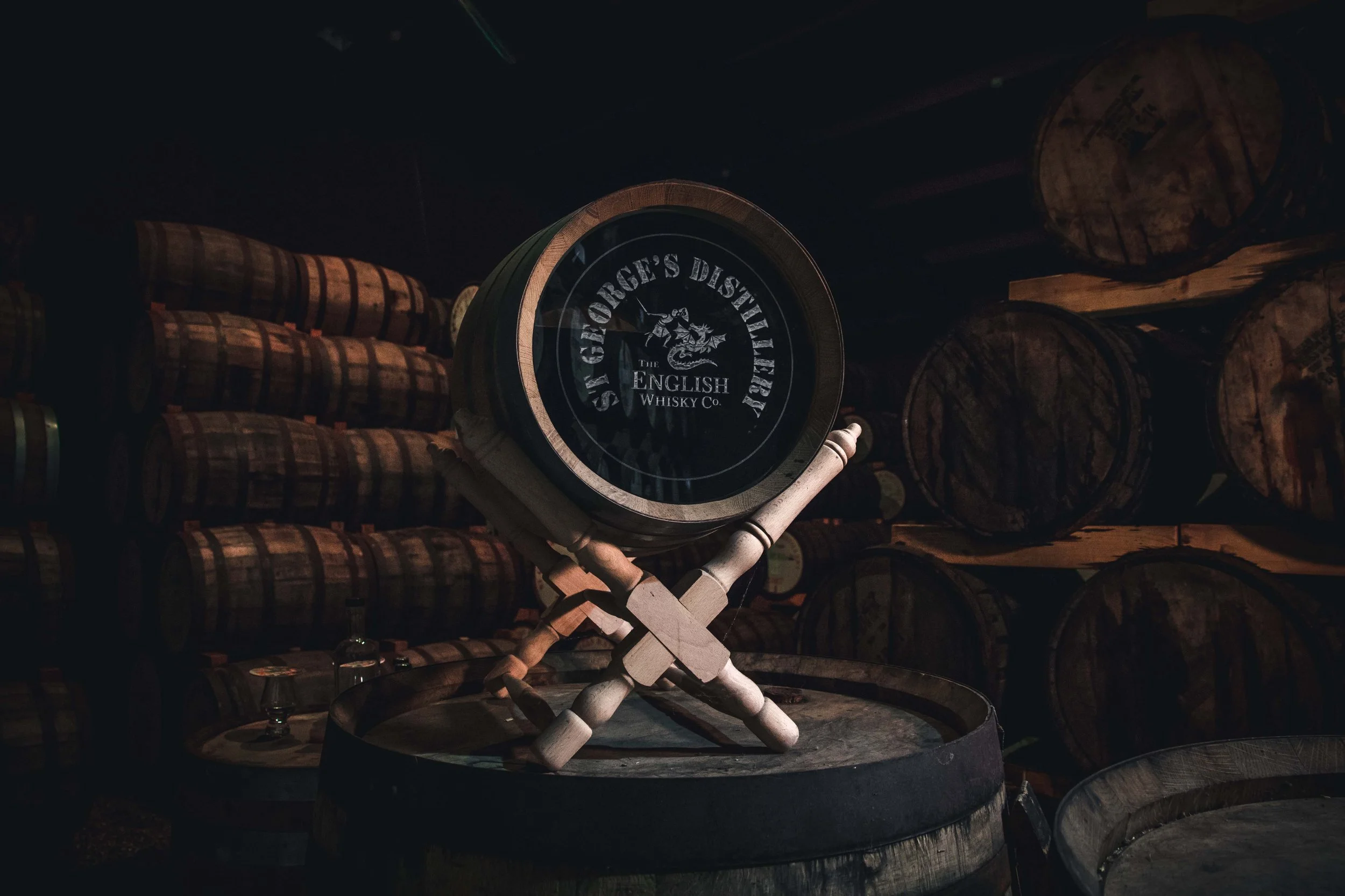 A wooden barrel with a display on top that reads 'St. George's Distillery, The English Whisky Co.', with stacked barrels in the background.