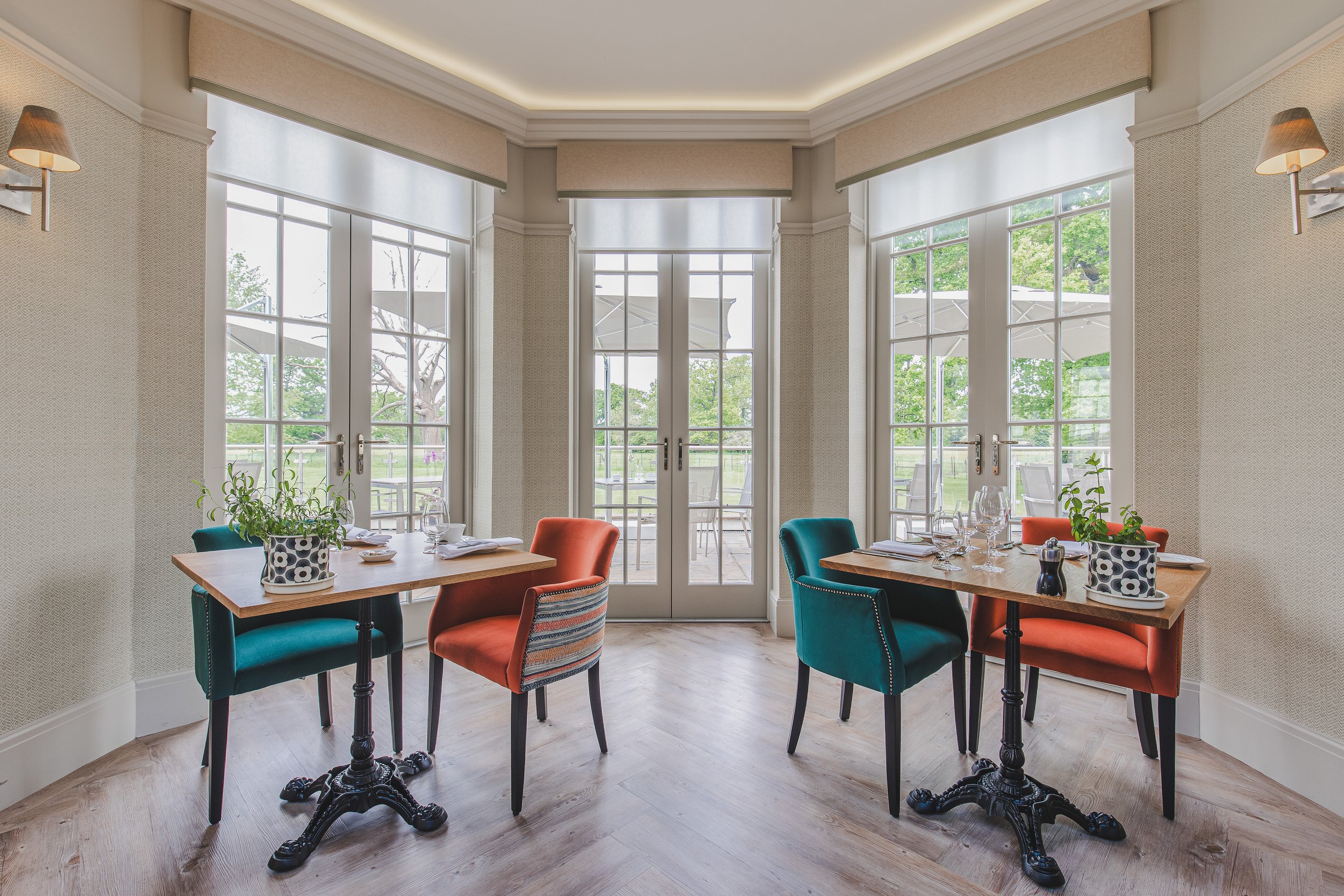Dining area with two tables set for two, placed near large windows with views of outdoor patio and greenery.