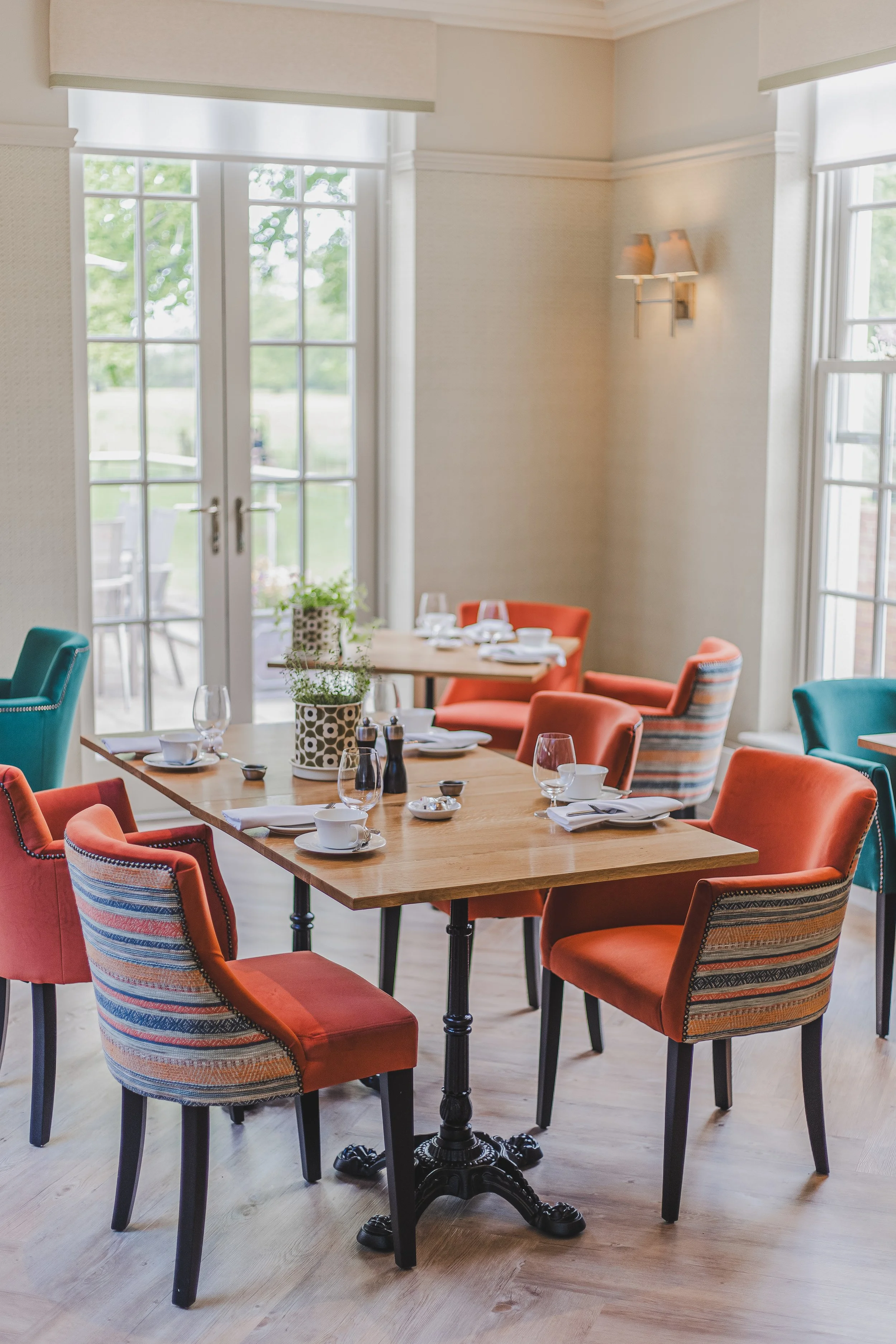 A dining room with wooden table and colorful upholstered chairs, set with cups, glasses, salt and pepper shakers, and flowers, next to glass doors leading to an outdoor patio.