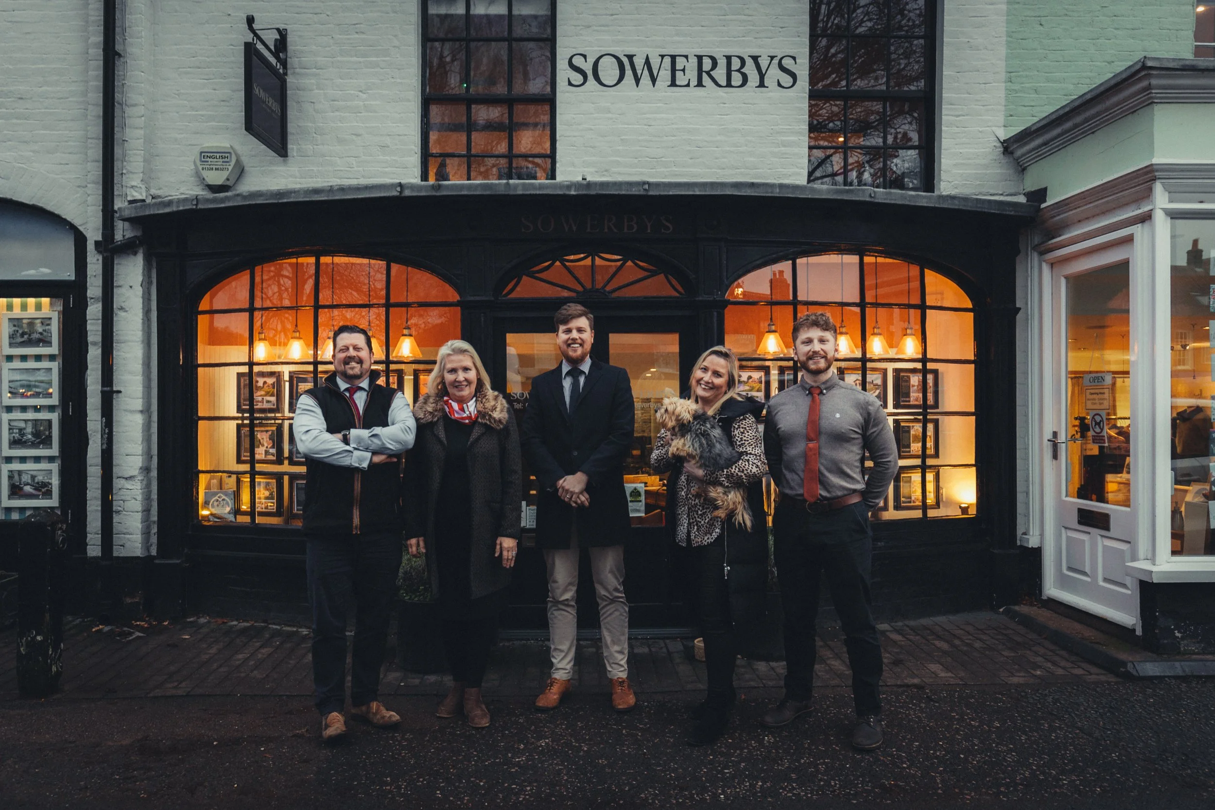 A group of five people standing outside a building named Sowerbys, smiling for a photo, with a woman holding a small dog, during evening hours.