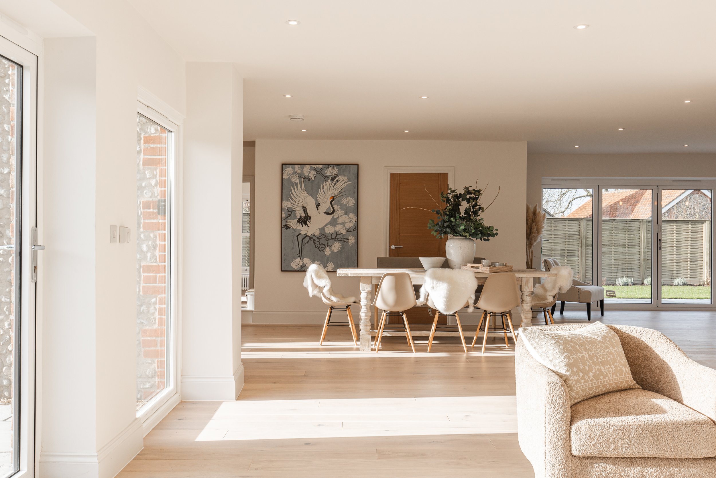 Bright, modern living room with large windows, a beige sofa, and a wooden dining table with white and fur-covered chairs. A large painting of a crane is on the wall, and there is a sliding glass door leading to a backyard with a wooden fence.