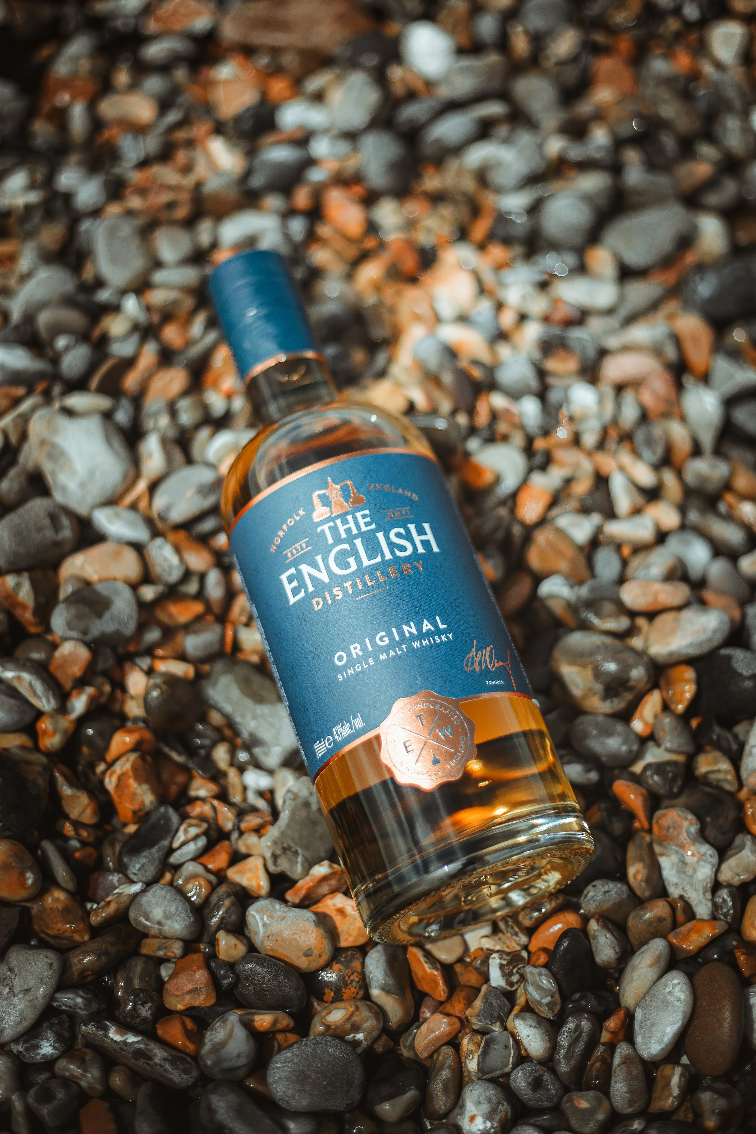 A bottle of The English Distillery Original single malt whisky lying on a bed of small, multicolored pebbles.