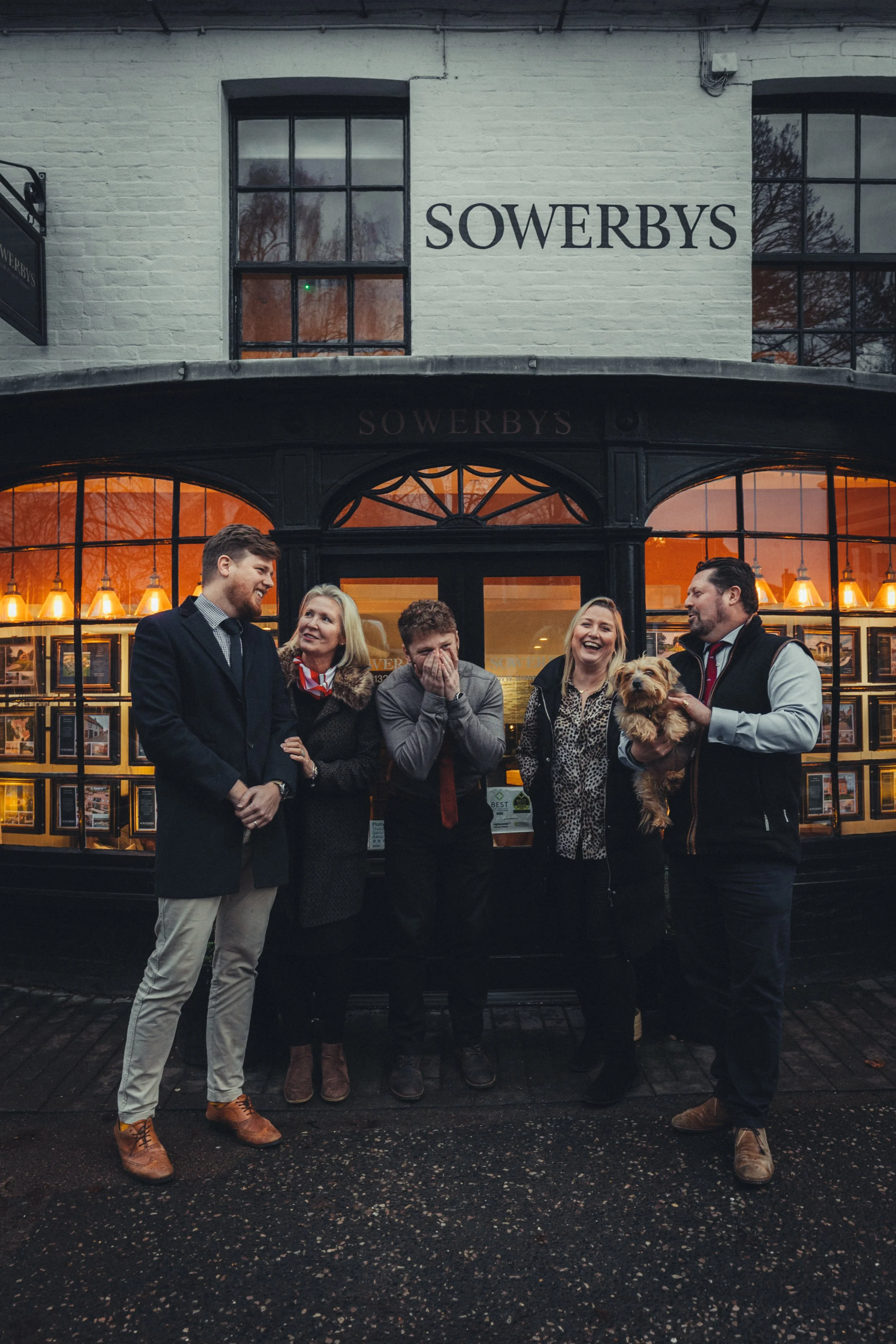 Group of five people standing outside Sowerbys real estate office, smiling and interacting, with a man holding a small dog, during early evening.