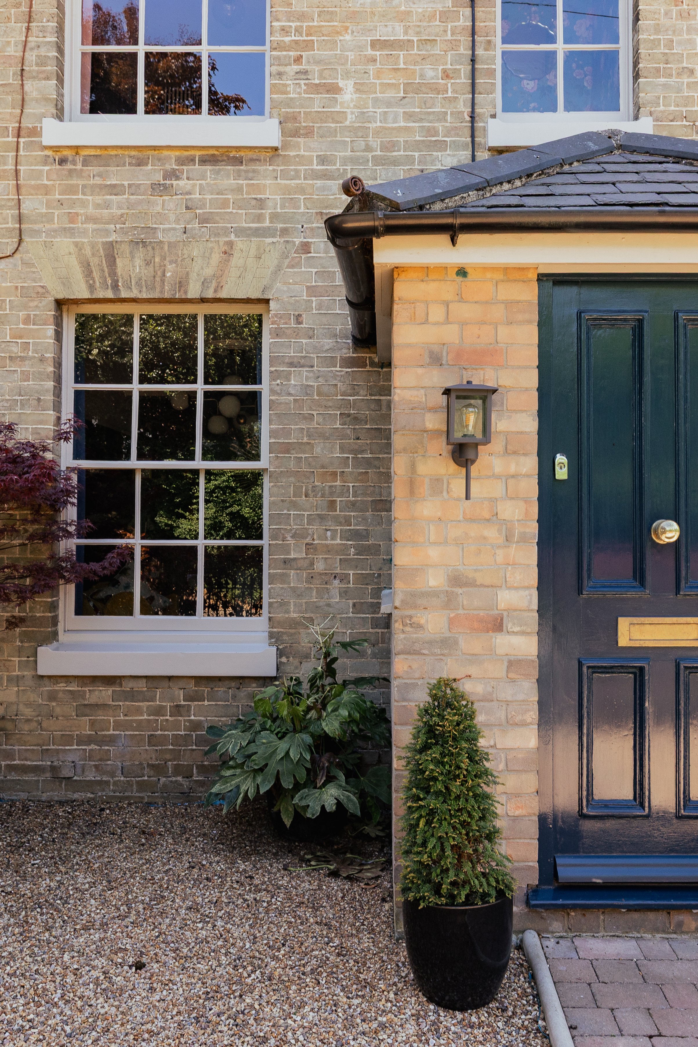 Close-up of a brick house exterior featuring a dark green front door, two large white-framed windows, a wall-mounted light, a small evergreen plant in a black pot, and a leafy shrub with large green leaves.