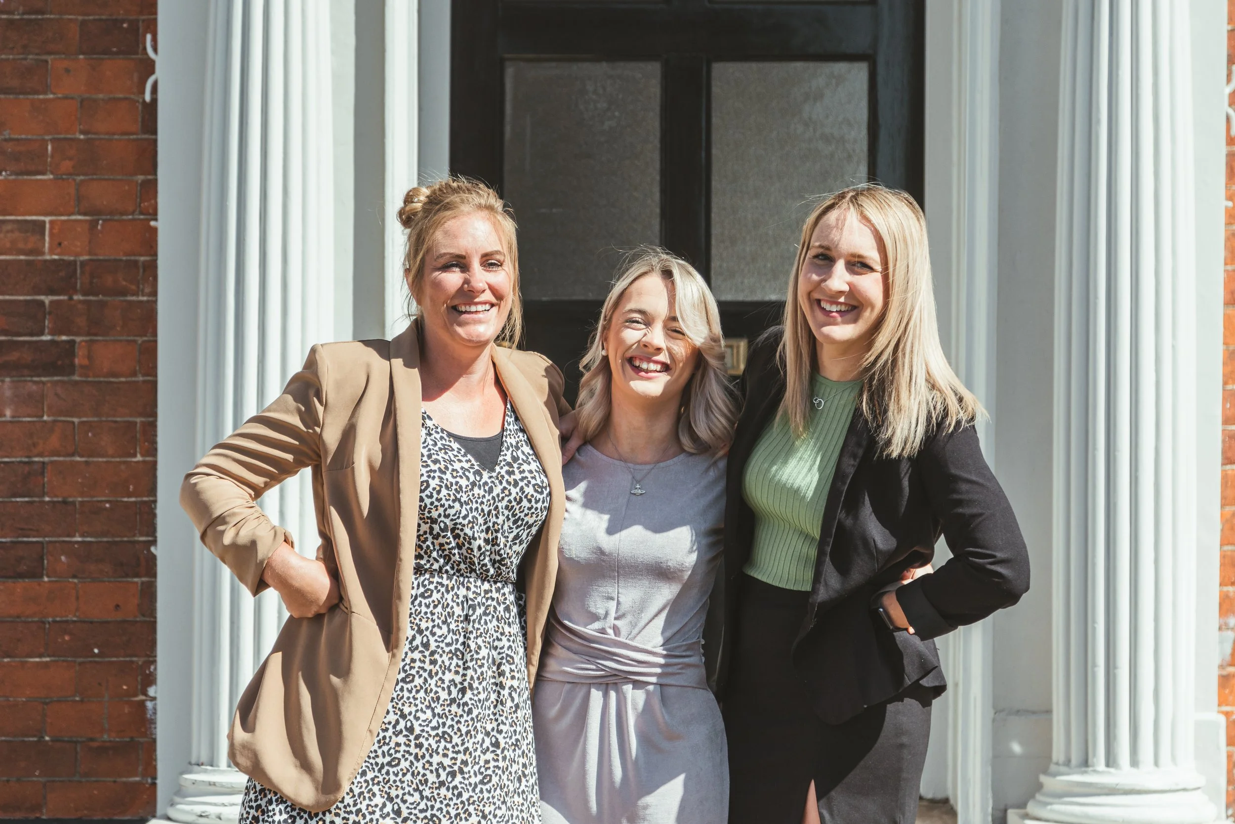 Three women standing outside in front of a house, smiling and posing together.