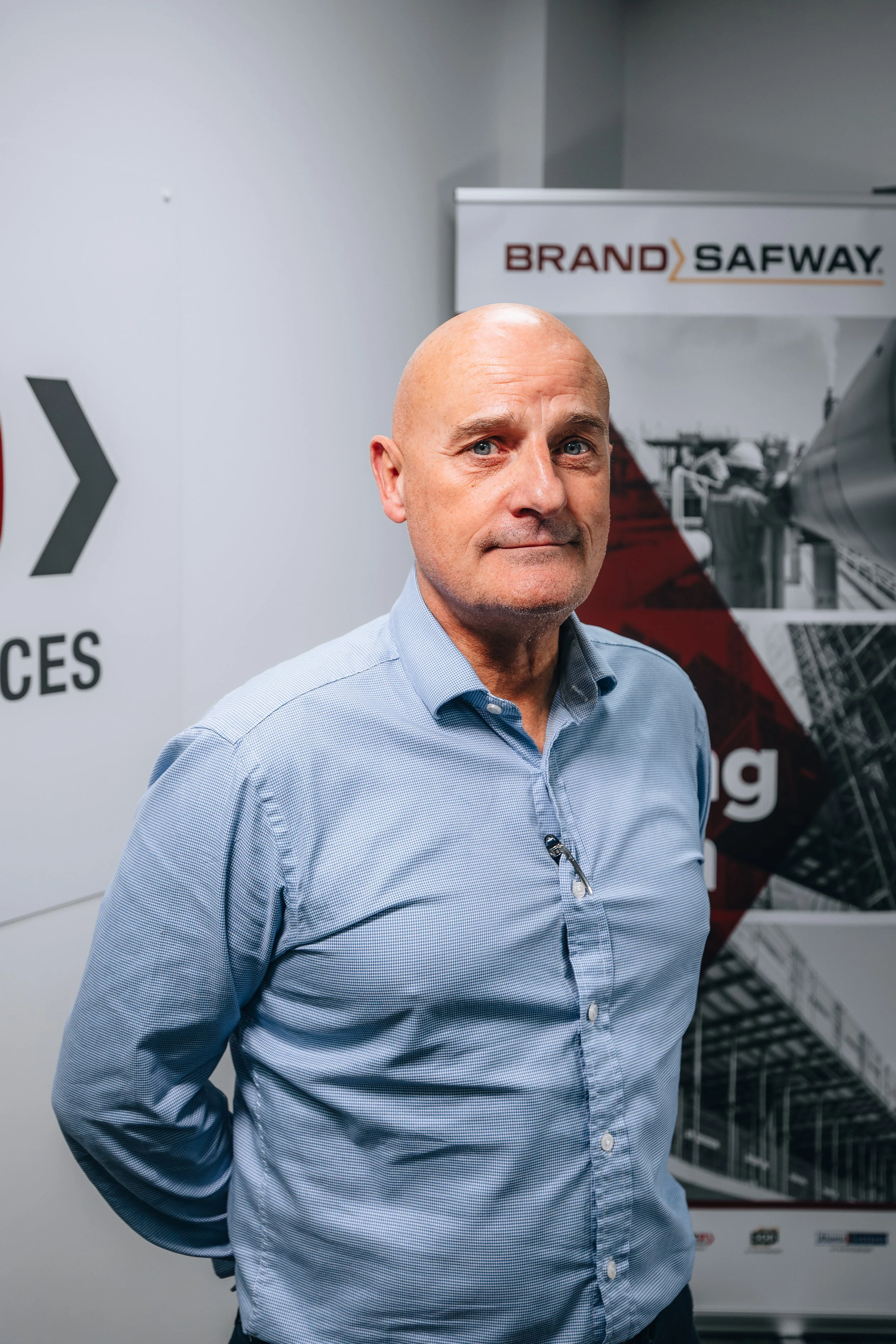 A middle-aged man in a light blue button-up shirt stands in front of an industrial-themed backdrop with the text 'BRAND SAFWAY' and a graphic of a construction site.