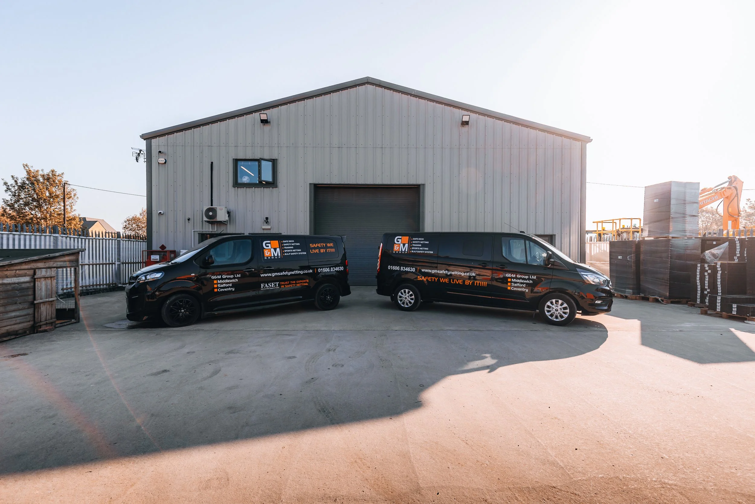 Two black company vans with branding parked in front of a large industrial warehouse with a roller door, surrounded by stacked pallets and construction equipment.