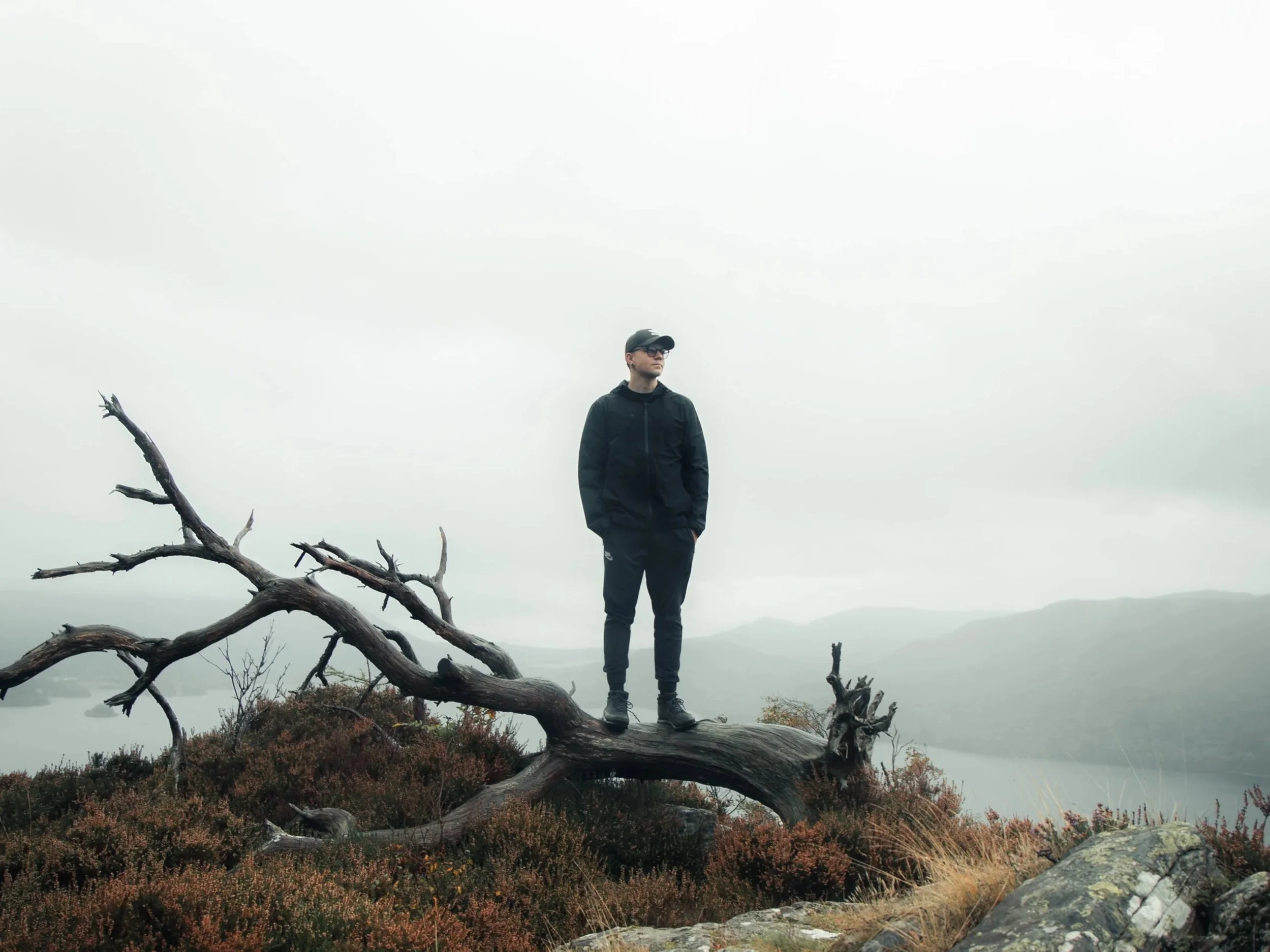A man standing on a fallen tree in a misty, mountainous landscape with a river in the background.