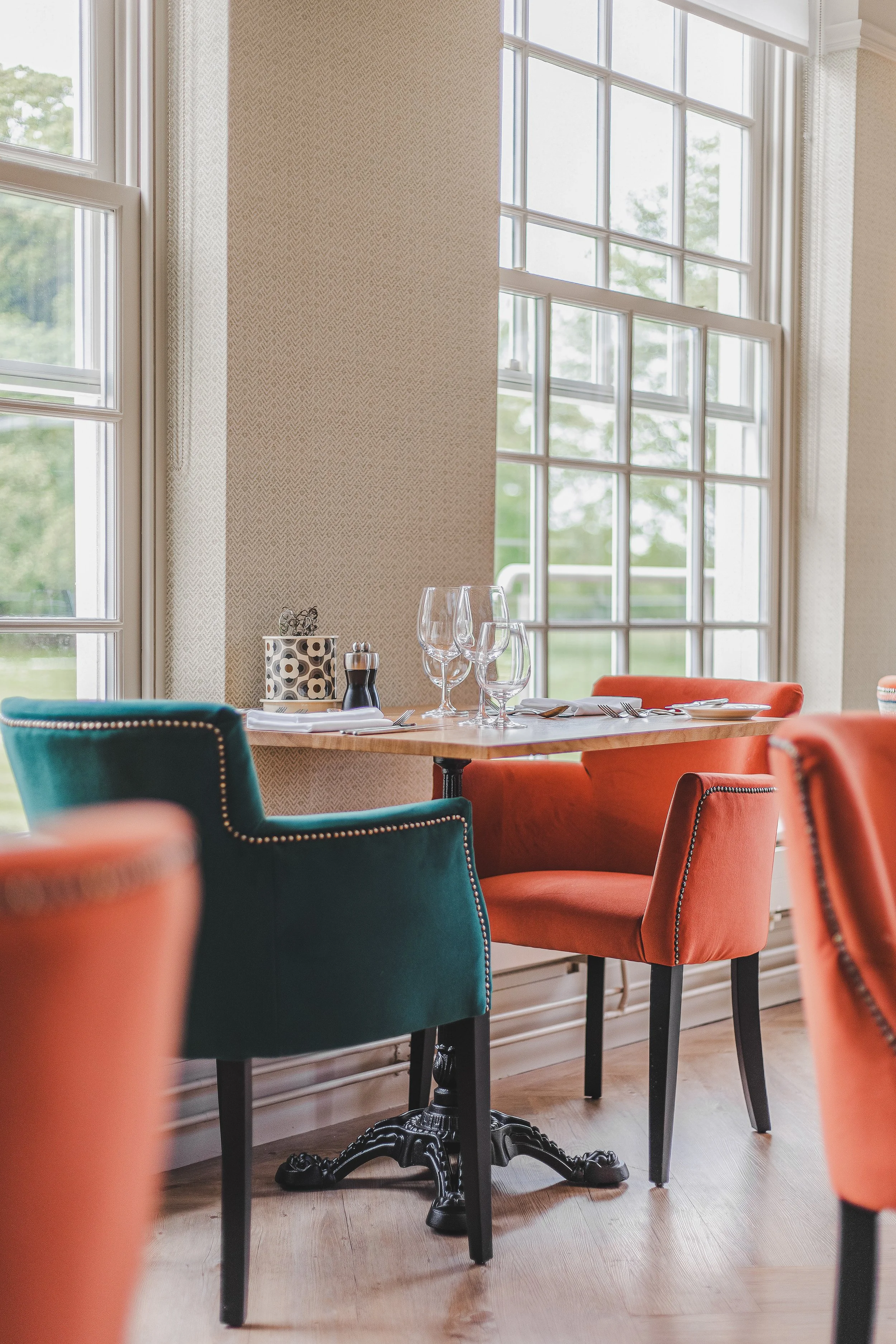 A dining table set near large windows, with green and orange upholstered chairs, wine glasses, and tableware.