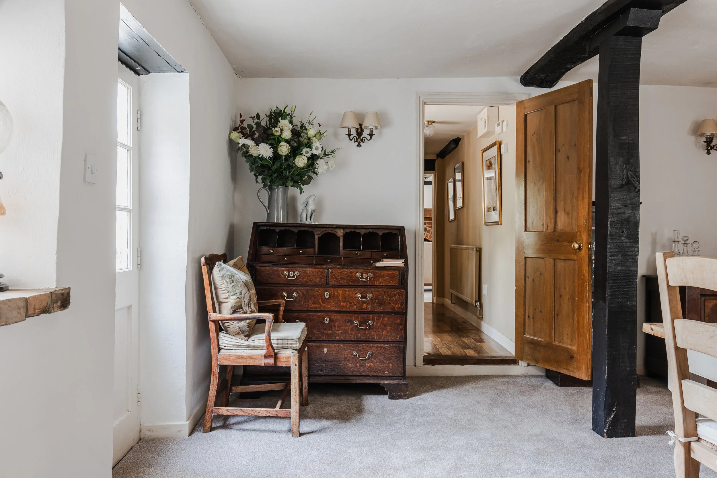 Interior of a cozy home with a wooden chest of drawers, a floral vase, a vintage chair with cushions, a partially open wooden door, a hallway with framed pictures, a chandelier, and a black wooden beam.