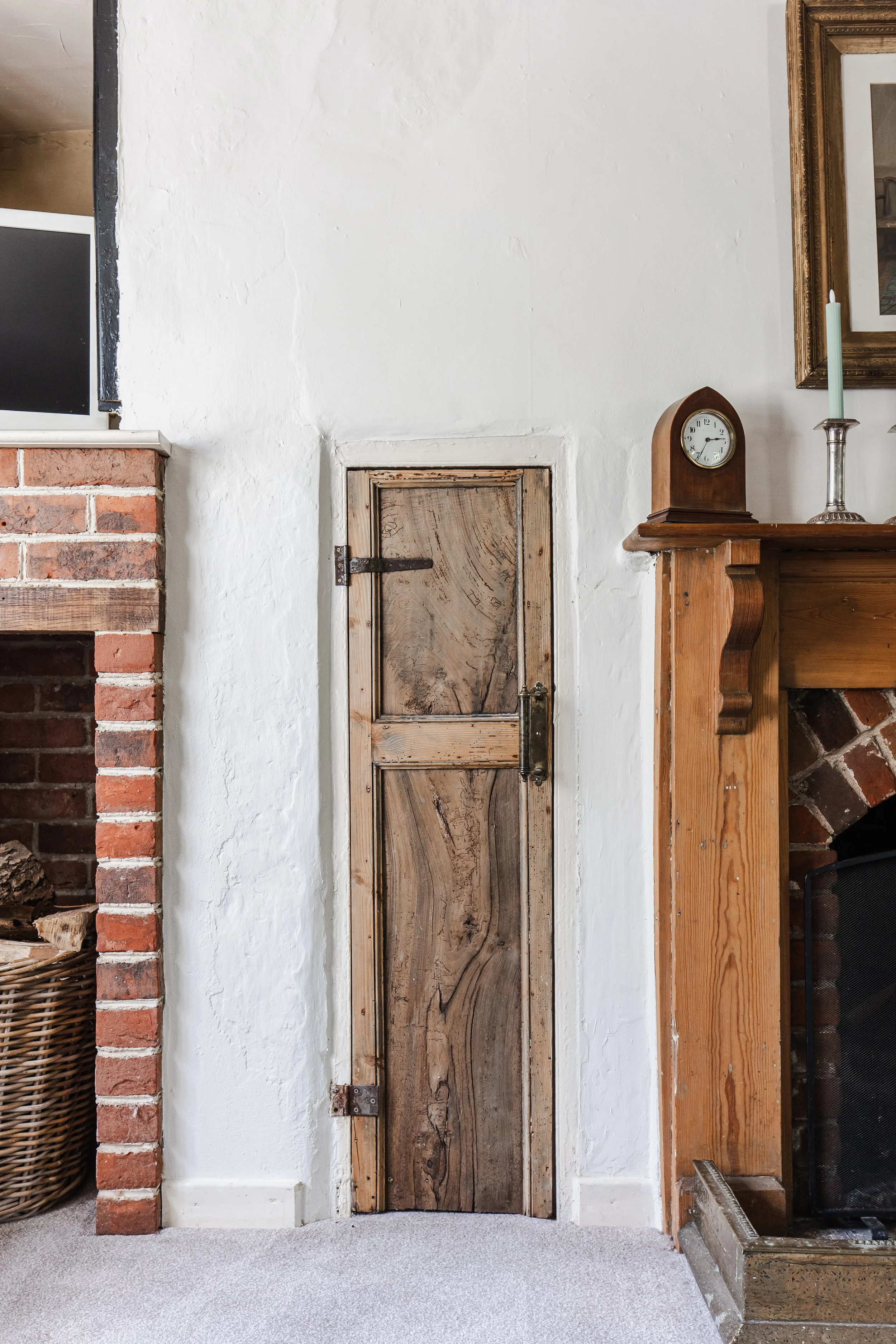 A small rustic wooden door set into a white textured wall, next to a brick fireplace with a basket of logs and a television, and above a wooden mantle with a clock and candlestick.