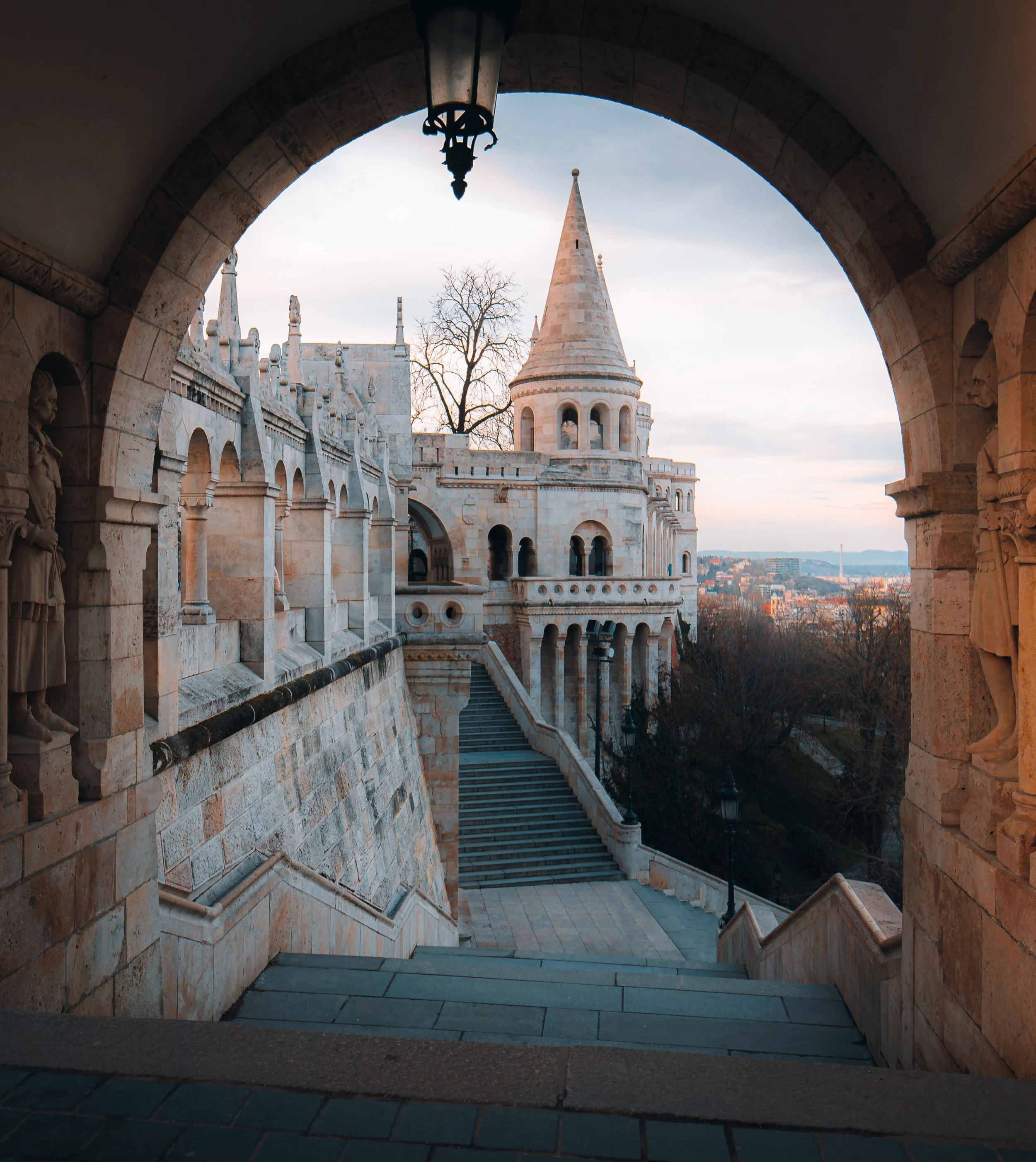 View of Fisherman's Bastion in Budapest, Hungary, through an arched stone gateway, showing stairs, turrets, and decorative architecture against a sunset sky.