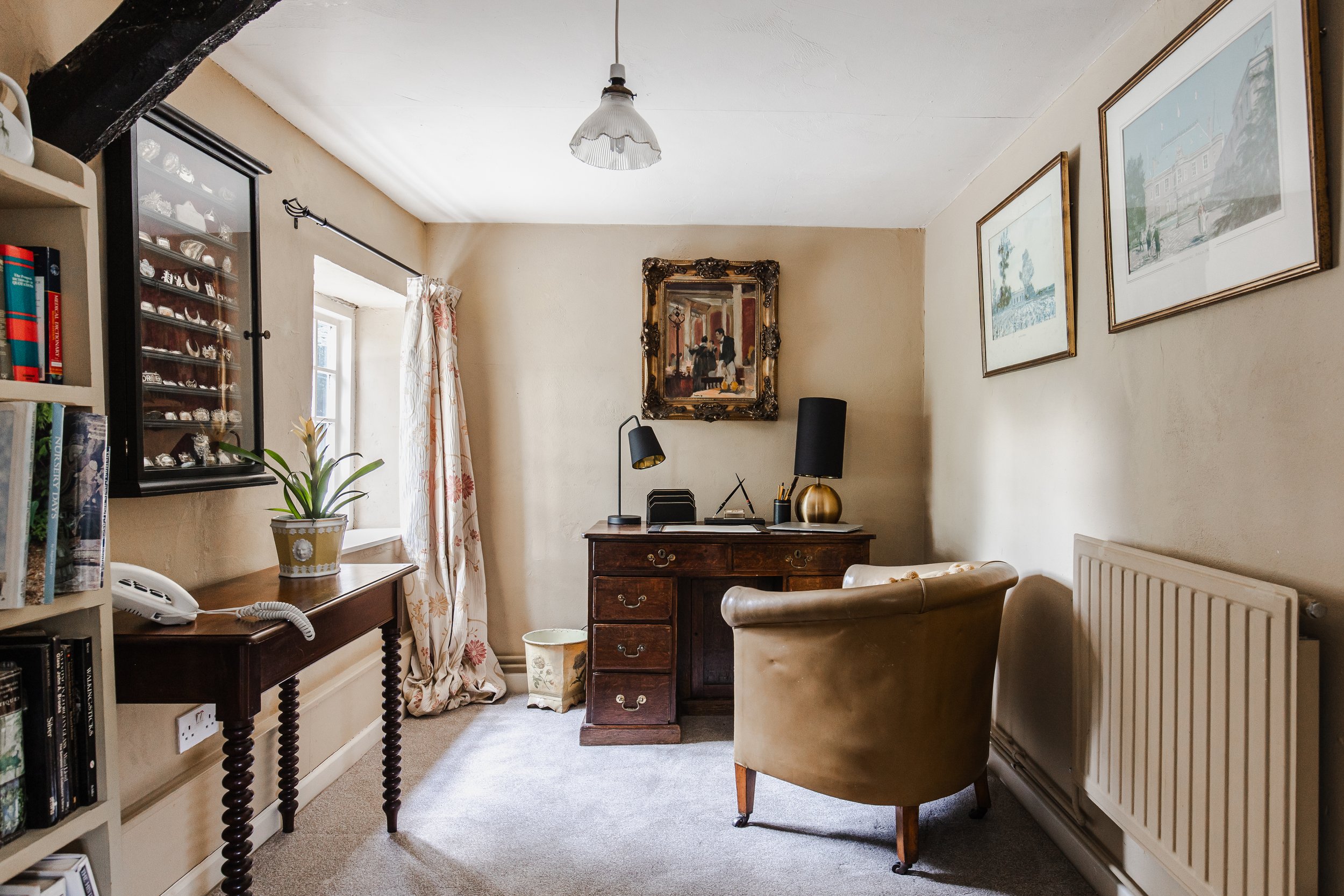 A cozy home office with a wooden desk, a beige armchair, paintings on the wall, a window with floral curtains, and a small cabinet with black lamps, a pencil holder, and decorative items.