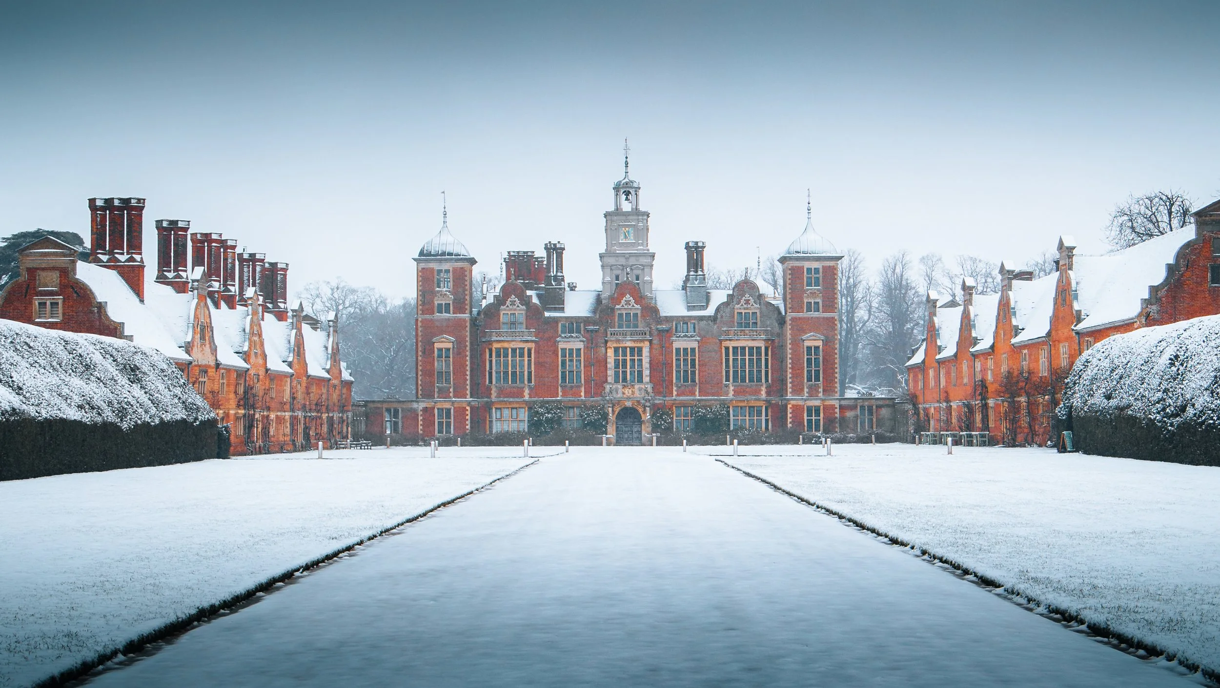 Large red brick castle with multiple turrets and a tall central clock tower covered in snow, surrounded by snowy grounds and trees.