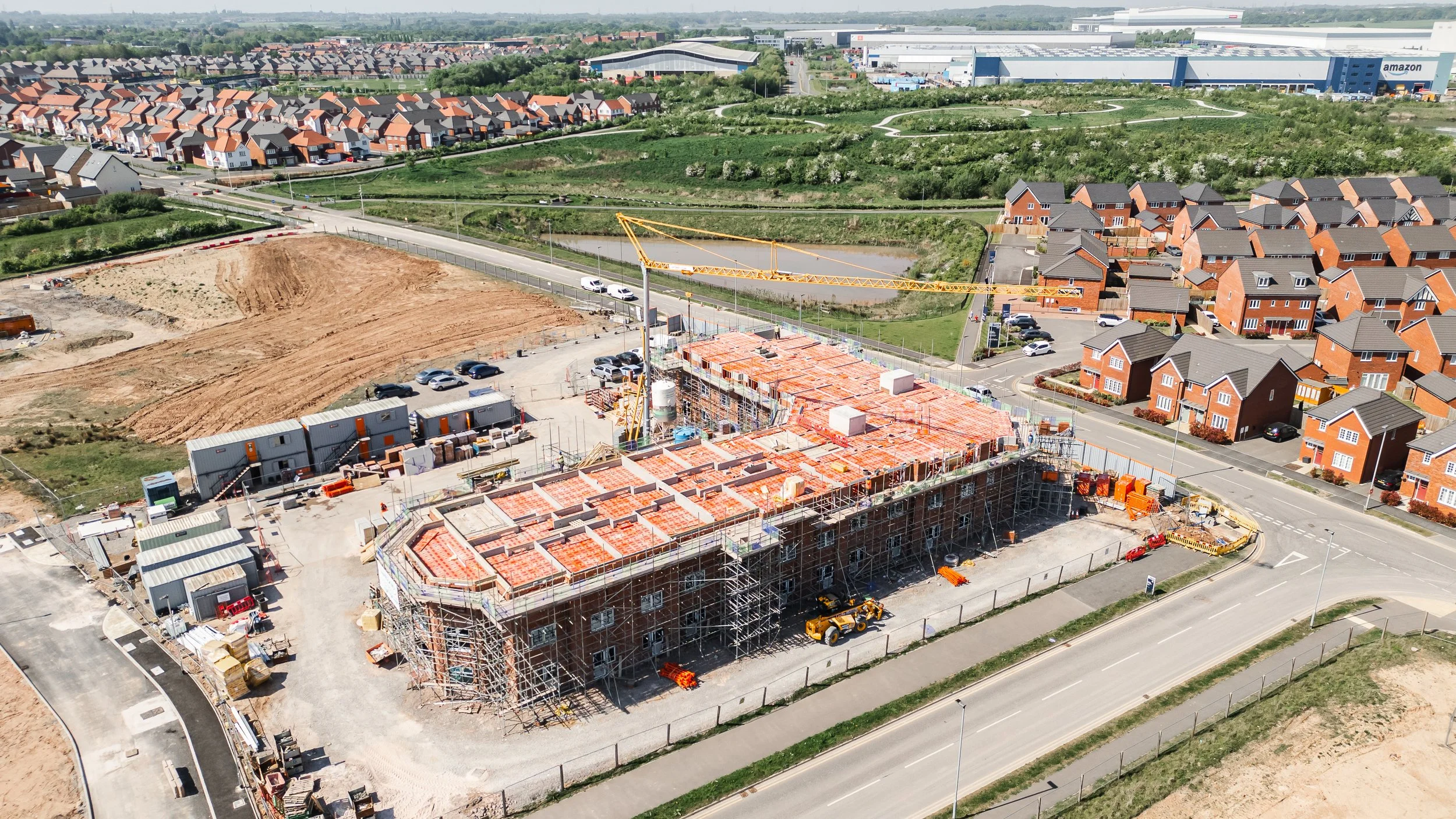 Aerial view of a construction site with scaffolding and a crane, adjacent to a residential neighborhood with new houses, near a commercial area with Amazon buildings.