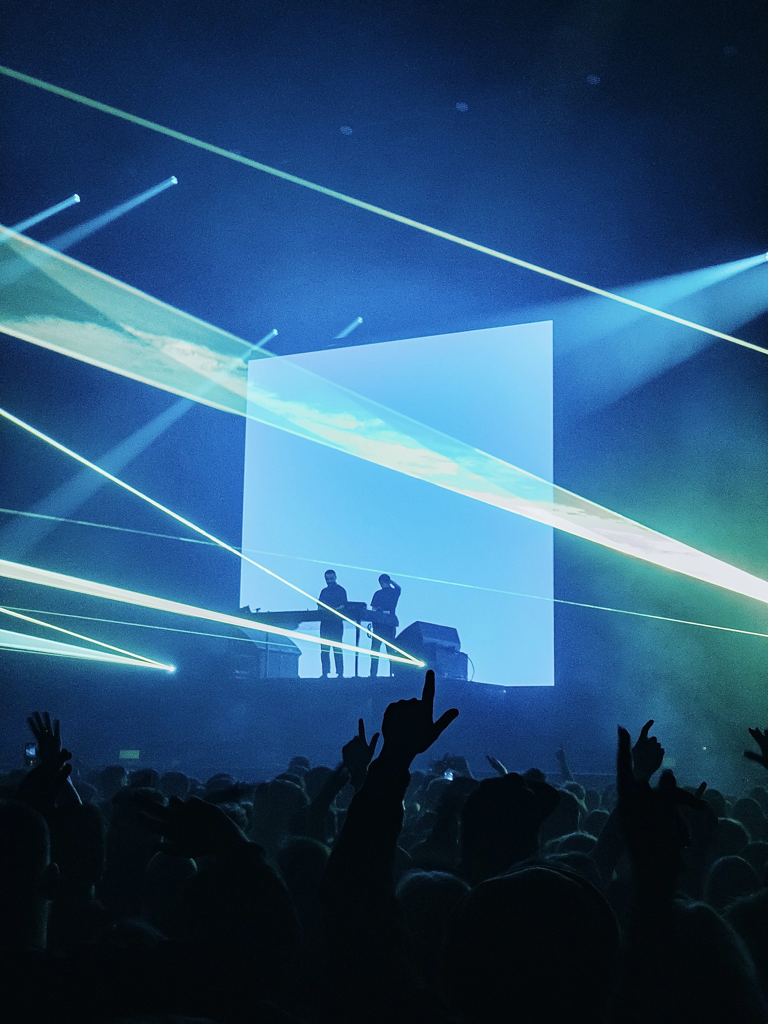 Concert scene with two performers on stage, bright laser lights, and silhouettes of audience members with some raising their hands.