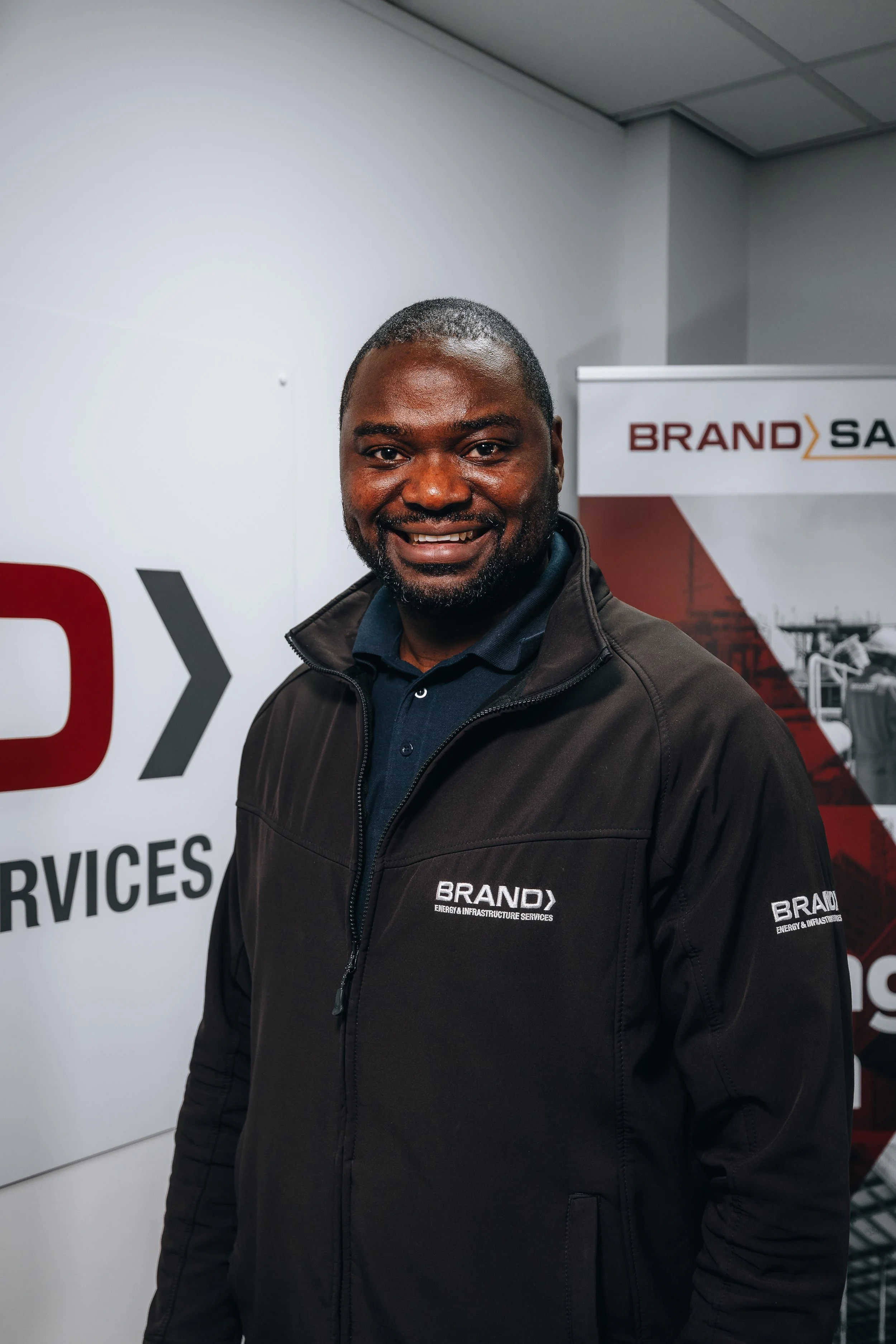 A man smiling in front of a branded backdrop, wearing a black jacket with the logo 'BRAND' and text 'Energy & Infrastructure Services'.