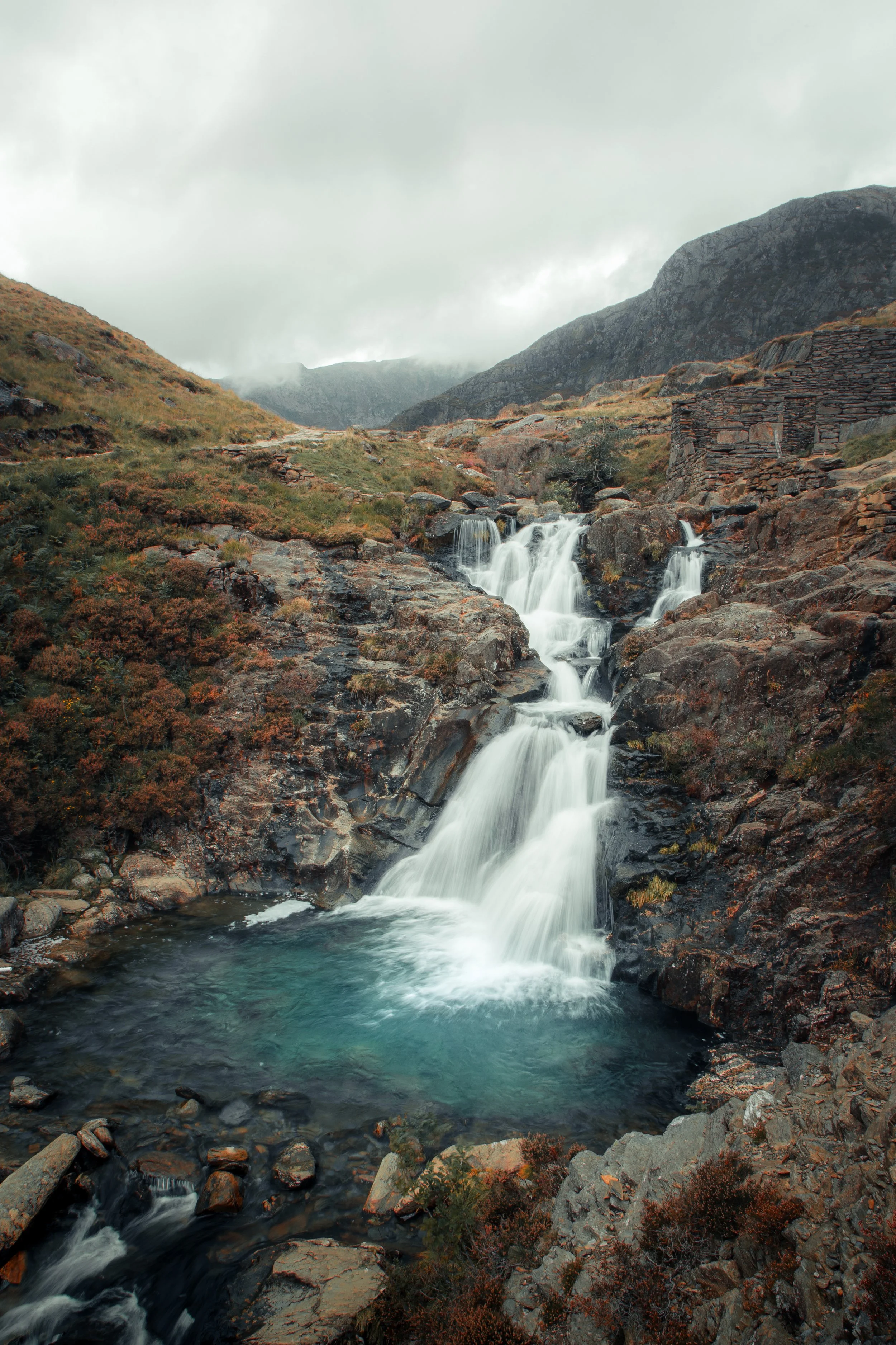 Scenic mountain landscape featuring a cascading waterfall flowing into a blue pool, surrounded by rocky terrain and misty mountains in the background.