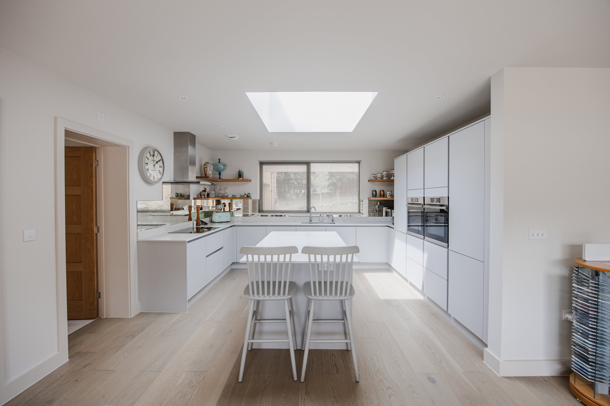 Modern white kitchen with a skylight, two white chairs at a white island, and built-in appliances, with wooden accents and decorative items on open shelves.