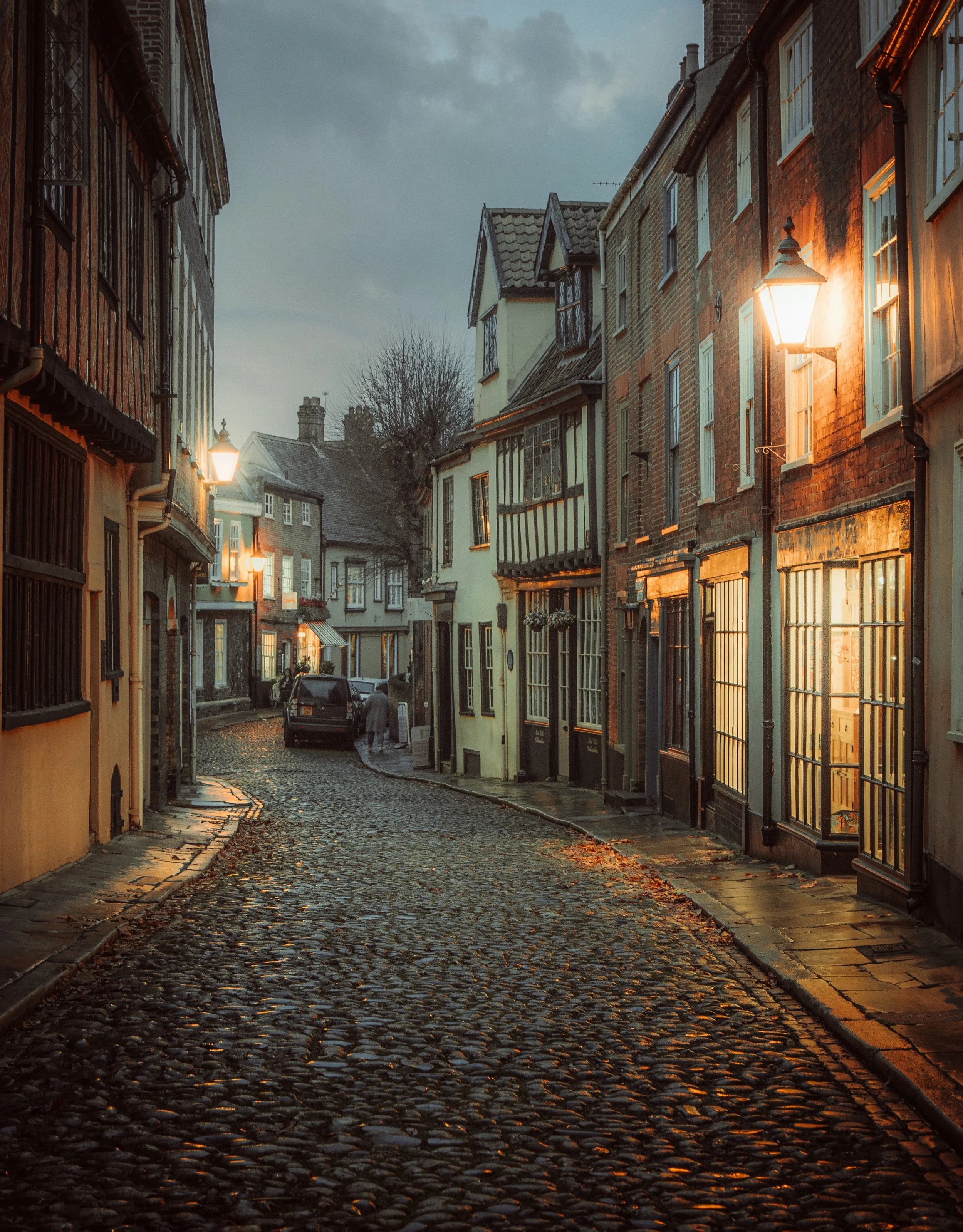 A cobblestone street in a historic European town during evening, lined with old brick and timber-framed buildings illuminated by street lamps.